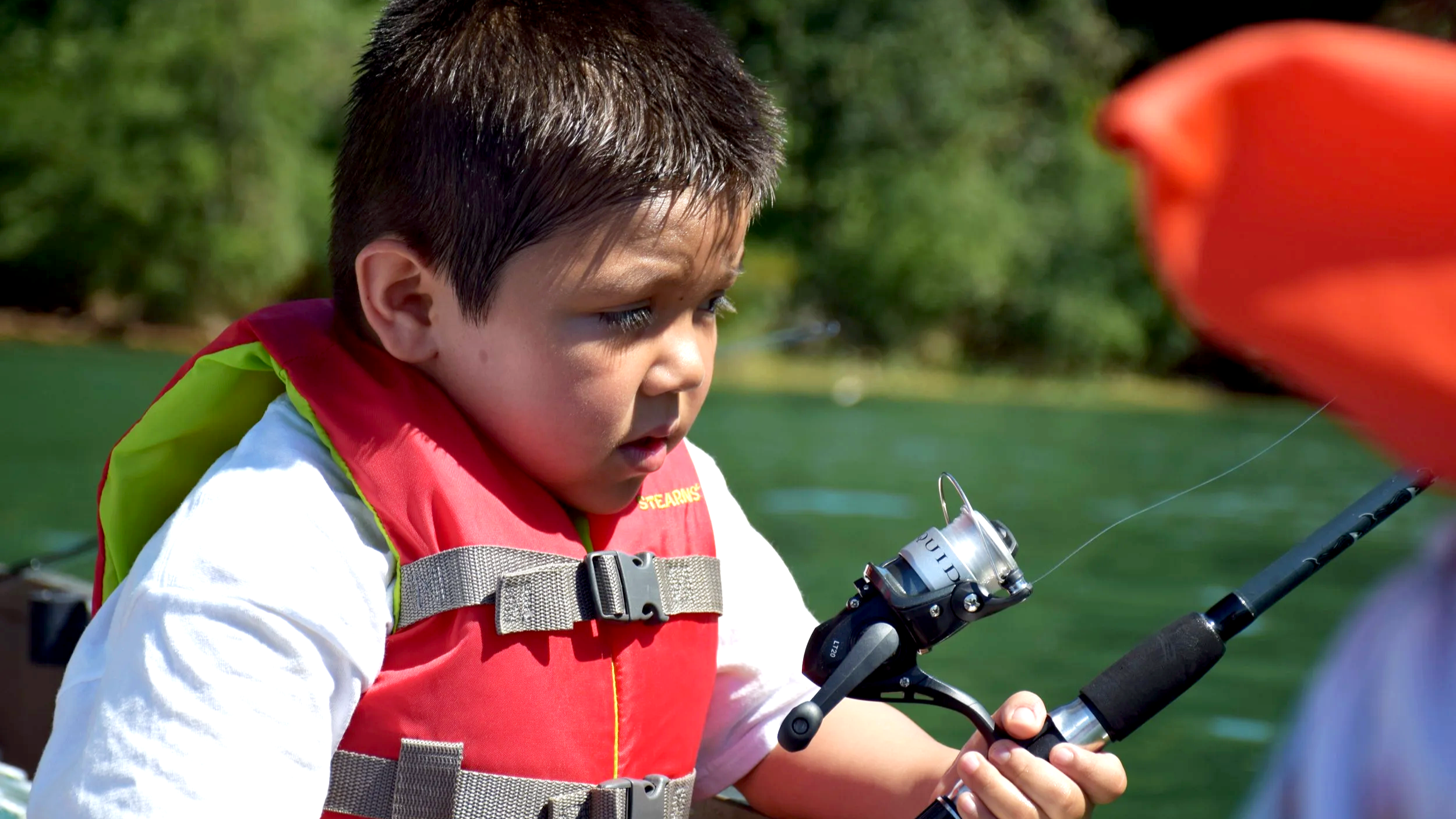 Young boy with dark hair wearing a red life vest and a white shirt, holding a fishing rod on a boat, looking attentively at the water.