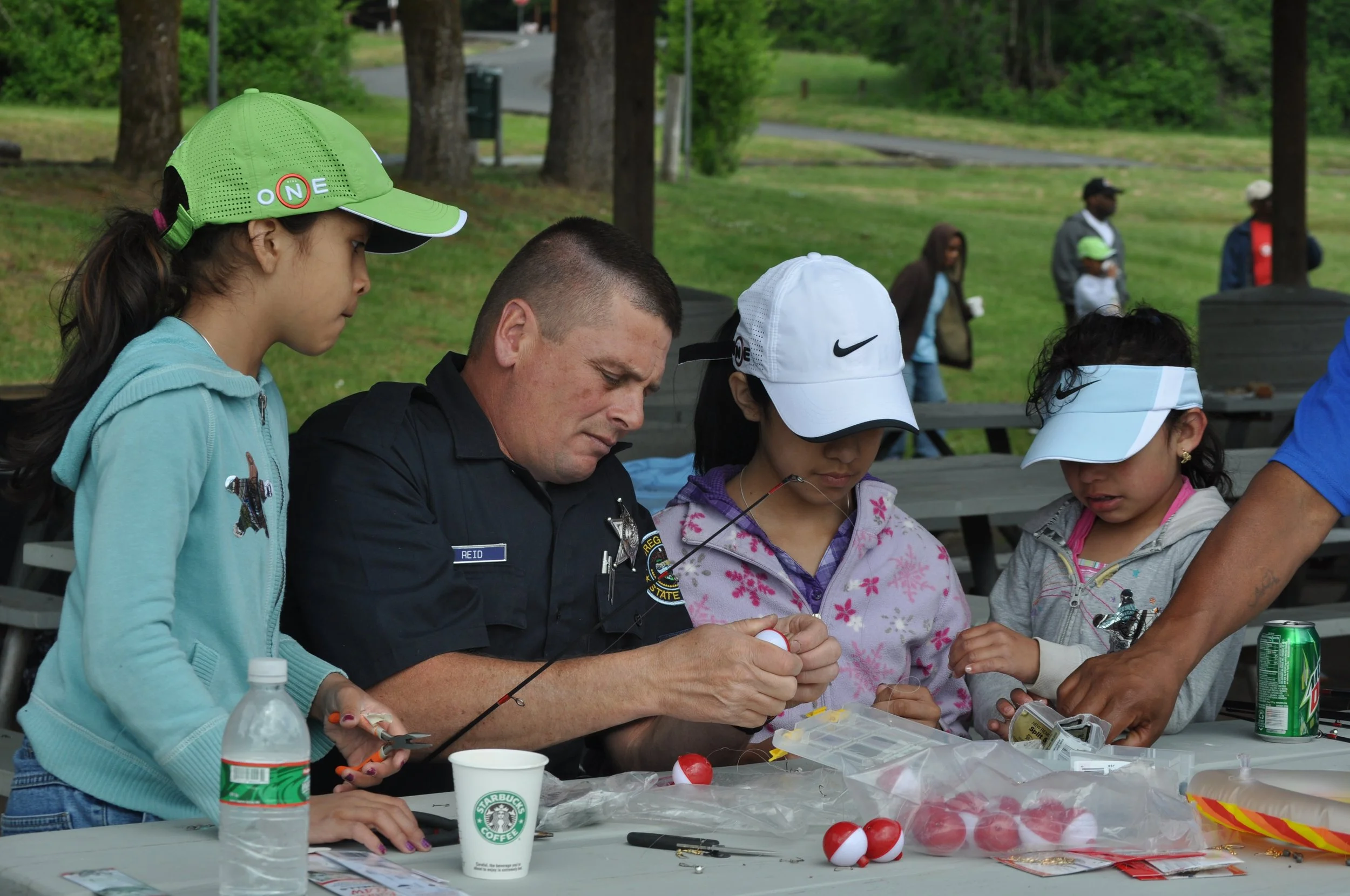 A police officer and three children gathered around a table outdoors, fishing gear and bait on the table, with people walking in the background.