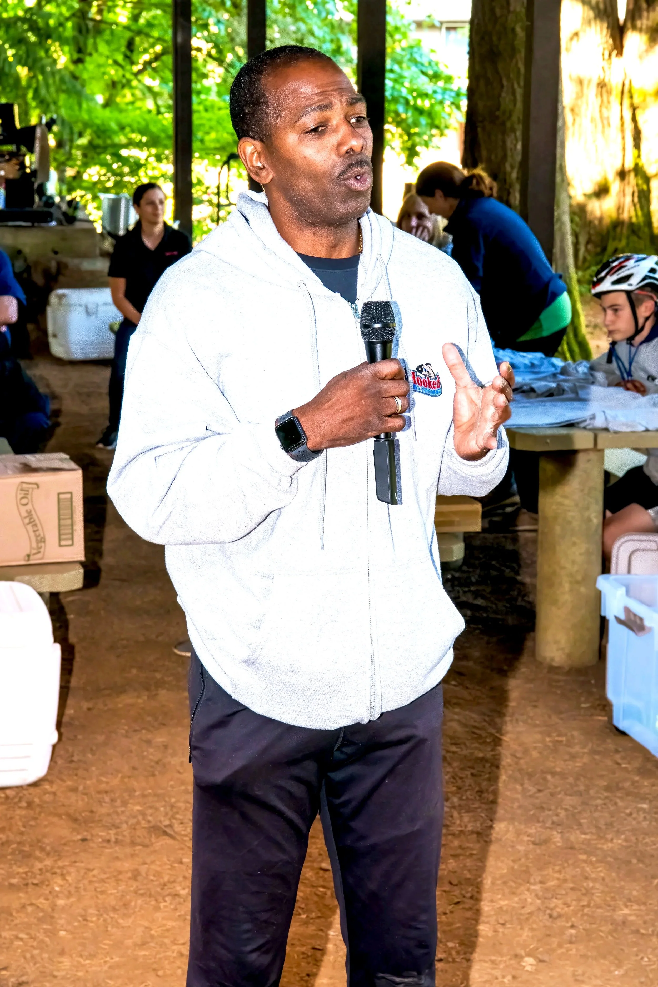 A man holding a microphone, speaking at an outdoor event in a wooded area with people sitting and standing around.