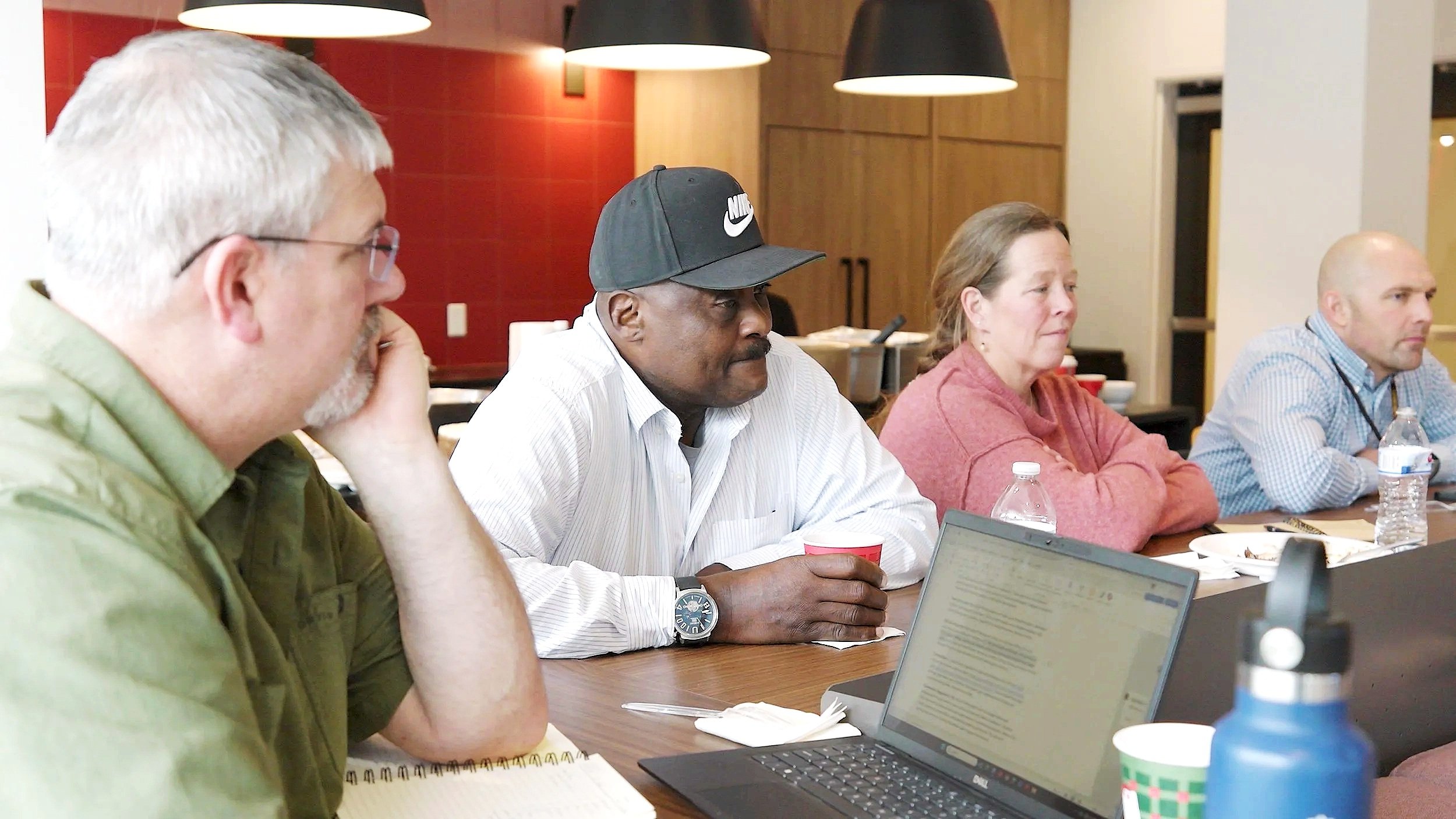 A group of four adults sitting at a conference table in a meeting room, listening attentively. The table has laptops, water bottles, and paper cups.