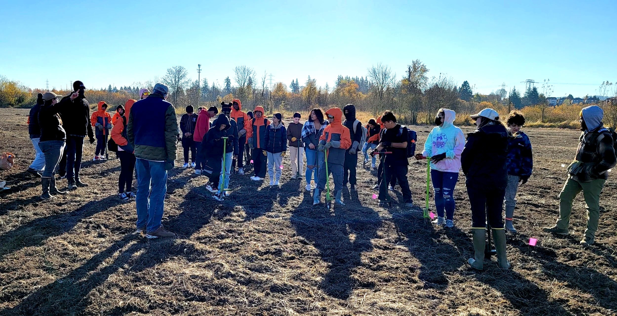 A group of people standing in a field during daytime, some holding tools, possibly participating in an outdoor activity or class, with trees and clear sky in the background.