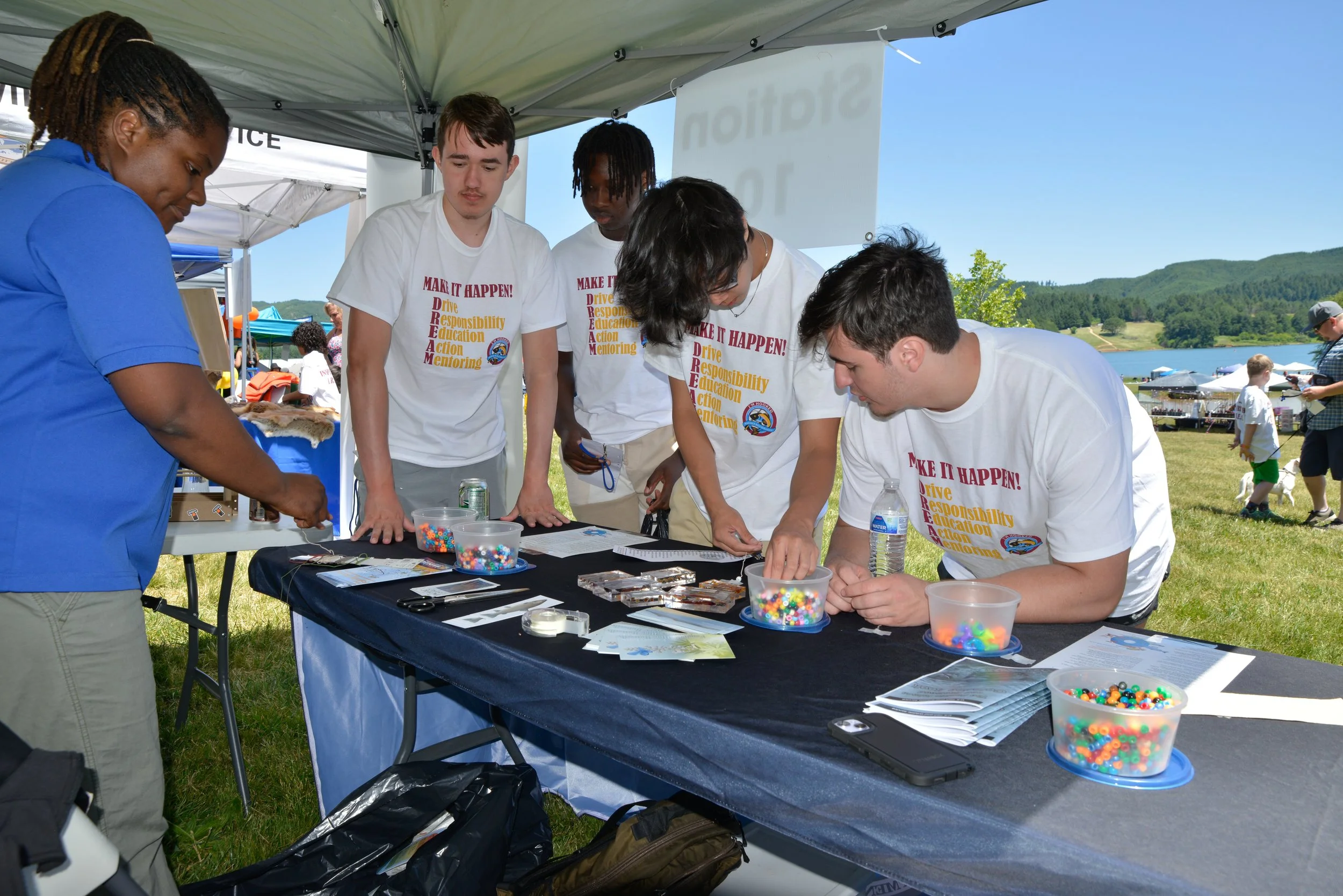 Group of young people at an outdoor booth, engaging in a craft activity with colorful beads, under a canopy with a lake and trees in the background.