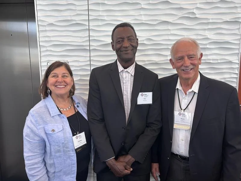 Three people smiling at a conference or professional event, wearing name badges, standing in front of a textured wall.