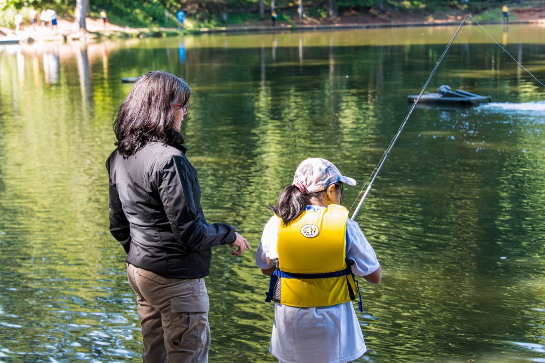 A woman and a girl fishing at a lake, with the girl wearing a yellow life jacket and a cap.