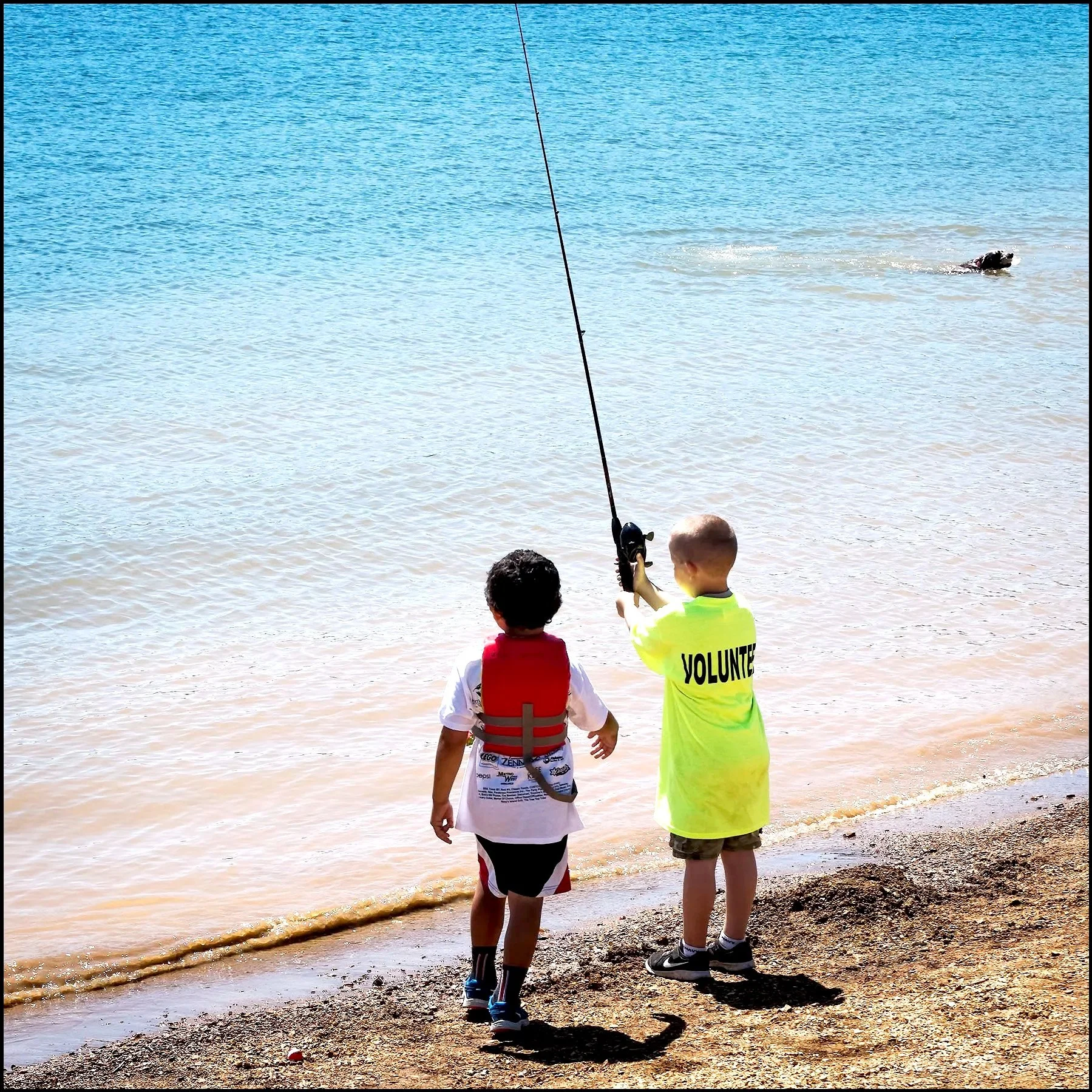 A boy in a yellow shirt labeled 'Volunteer' helping another boy in a white shirt with a red life jacket to fish at the water's edge on a beach.
