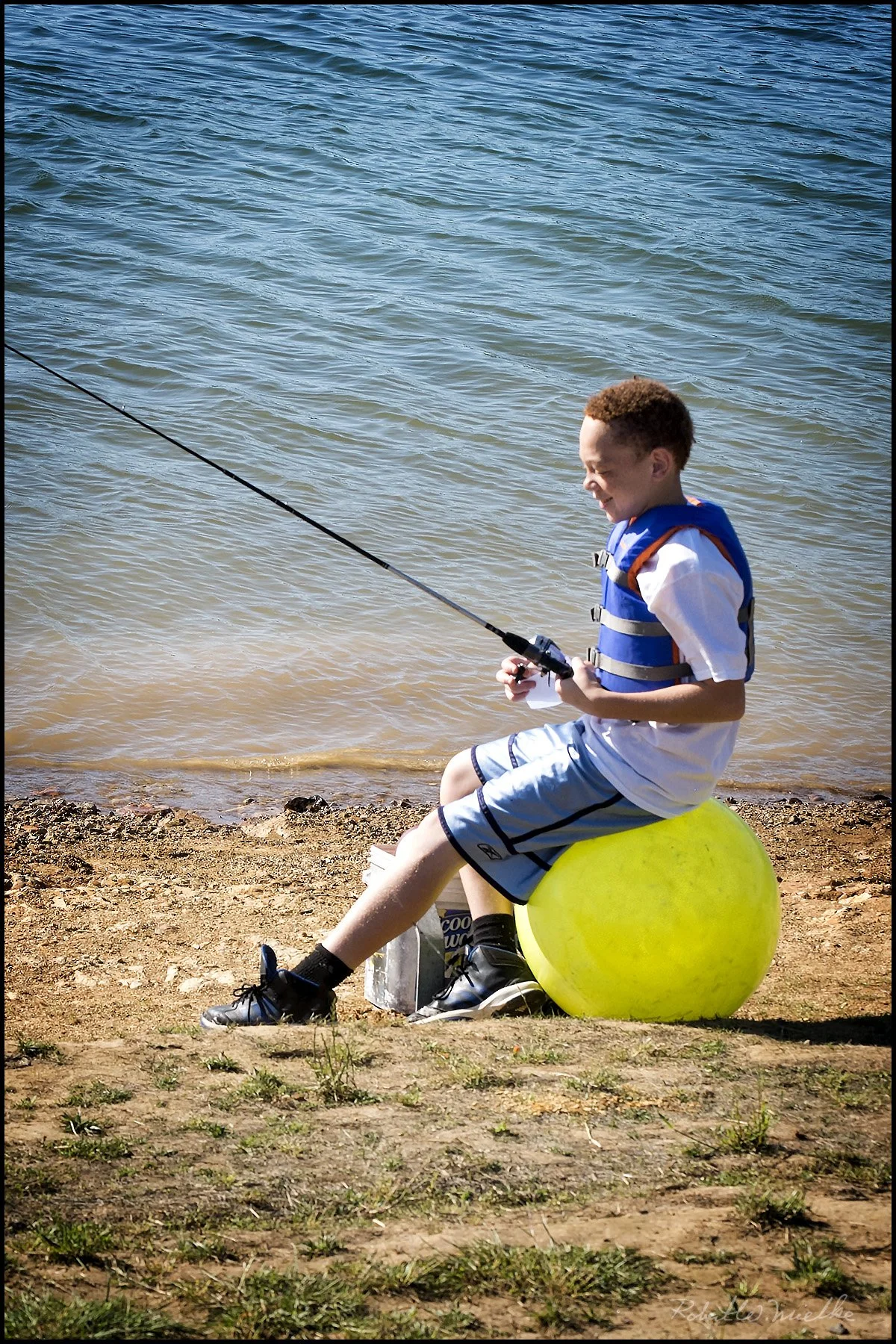 A boy sitting on a yellow buoy by the lake, fishing with a rod in hand, wearing a blue life jacket, white shirt, and shorts.