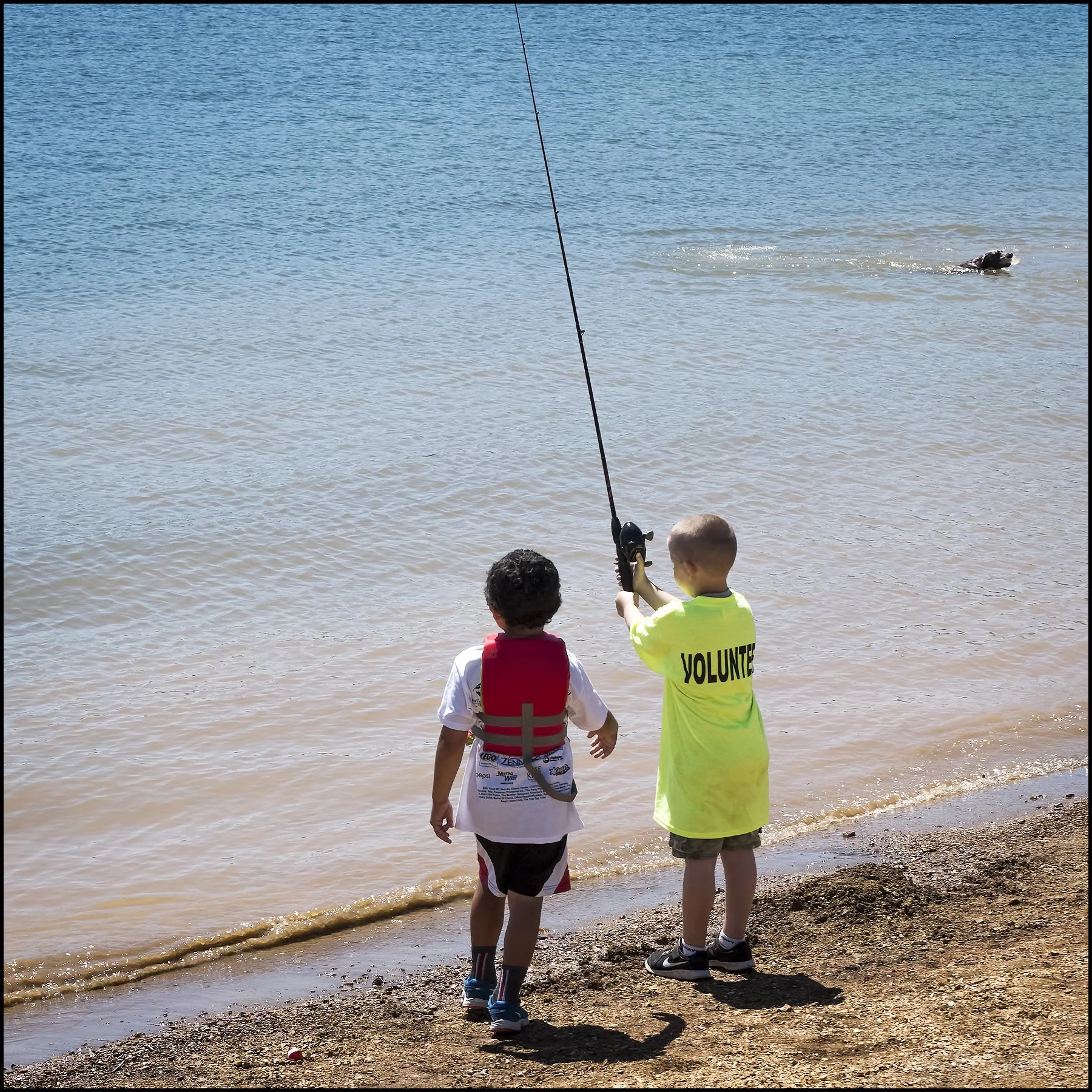 A young boy in a yellow volunteer t-shirt and shorts is fishing at the edge of a lake, while another child in a white t-shirt and shorts stands nearby observing. There is a dog swimming in the lake in the background.