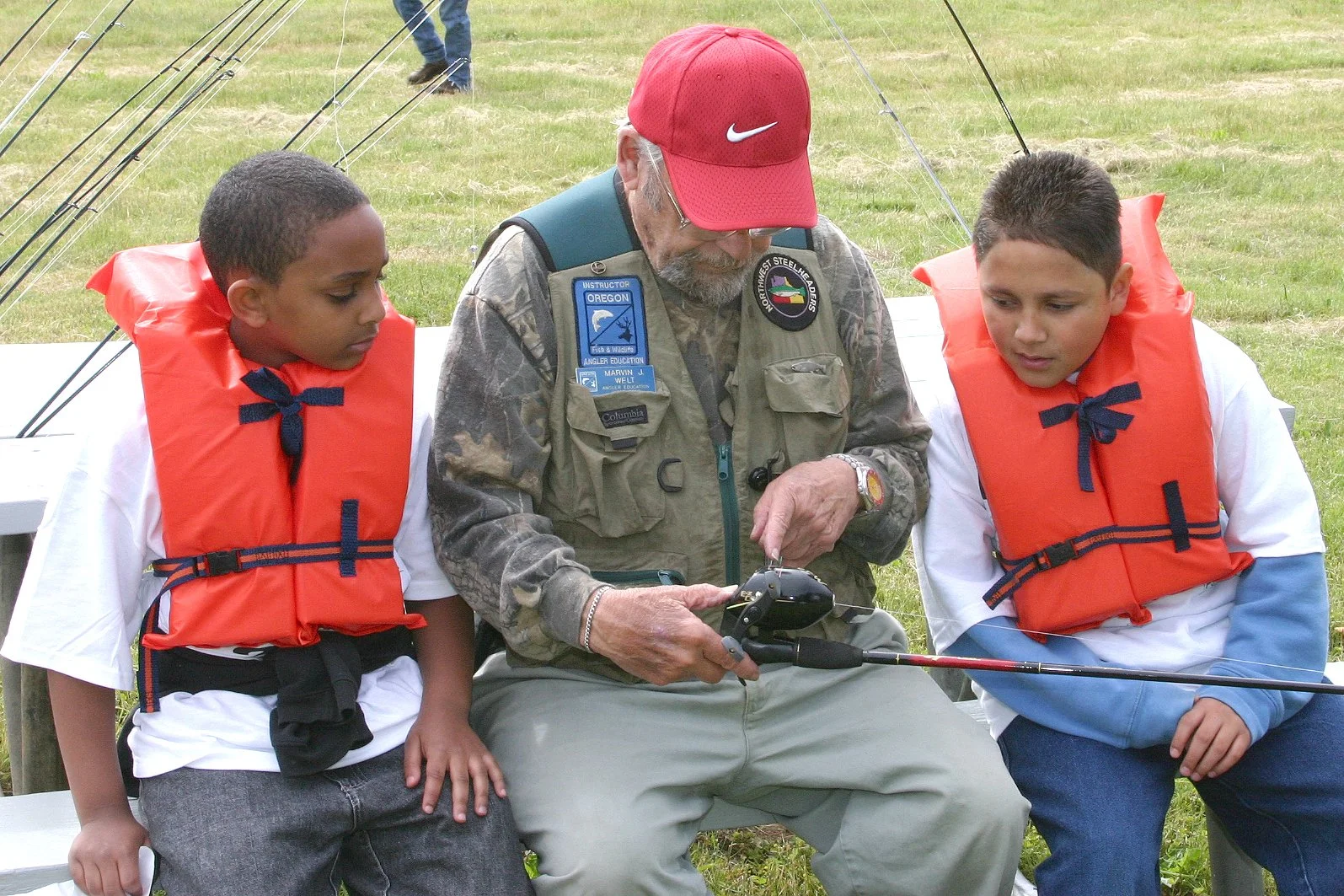 A man with gray hair and glasses, wearing a red Nike cap, camouflage jacket with several patches, and a watch, showing a fish fishing device to two young boys sitting on a bench, all outdoors in a grassy area.