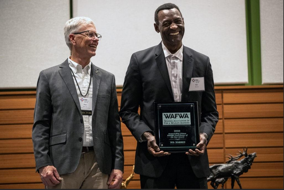 Two men in suits standing on a stage, one holding an award plaque, with animal statues and a green and wooden backdrop behind them.