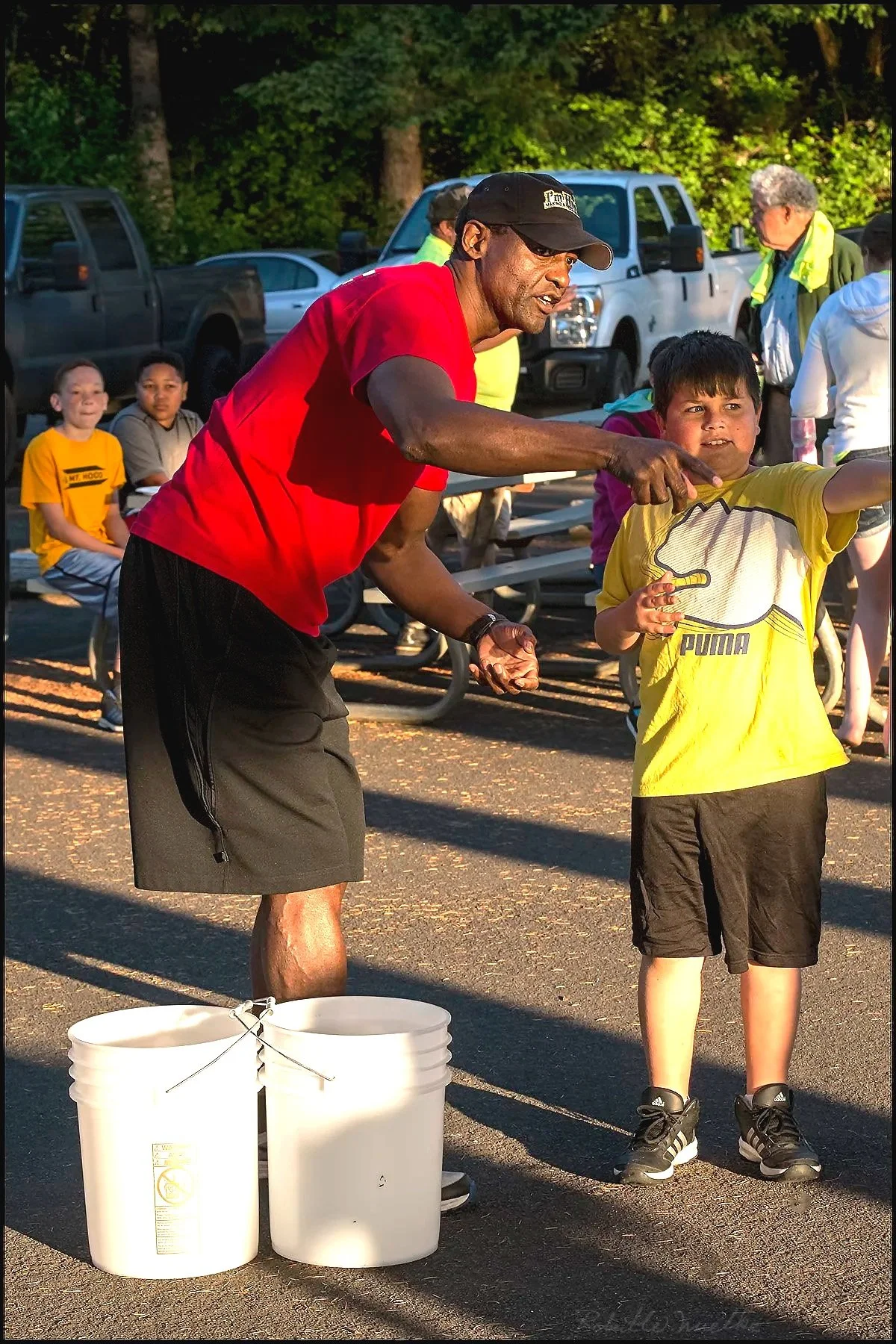 A man in a red shirt and black shorts helping a young boy in a yellow shirt at an outdoor event, with children and adults sitting and standing nearby.