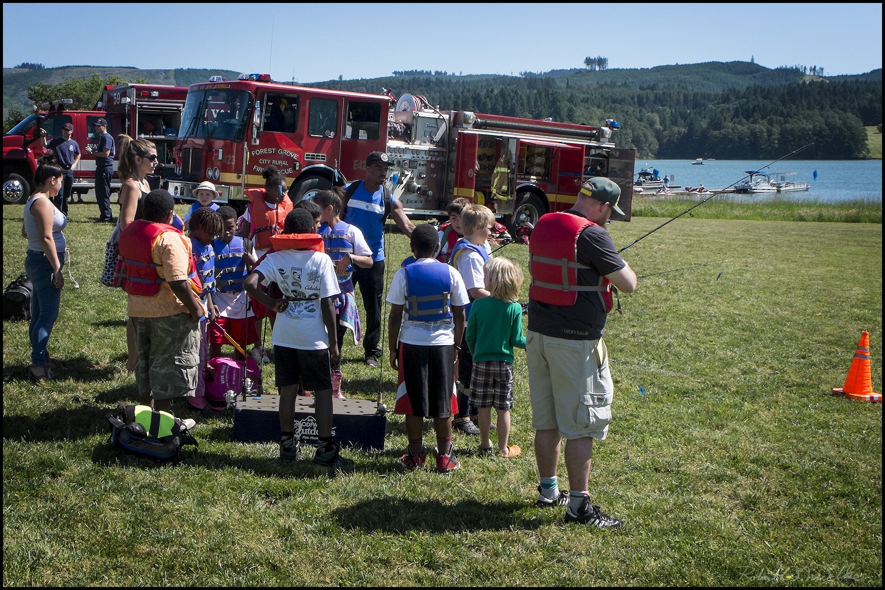 Children and adults gathered outdoors near a lake, where firefighters demonstrate fishing safety. Fire trucks are parked behind them, and some children are wearing life jackets while others hold fishing poles. The setting features green grass, water with boats, and forested hills in the background on a sunny day.
