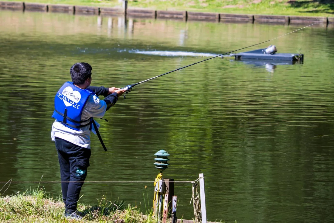 A boy fishing by a pond, wearing a life jacket and standing on the grassy bank, with a boat in the water in the background.
