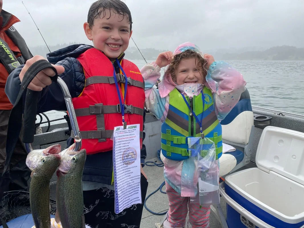 Two children on a boat, one boy holding two fish on a hook, and a girl smiling and adjusting her hood, wearing life jackets, with a cooler nearby and water in the background.