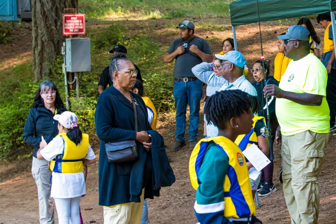 A group of diverse people, including children and adults, gathered outdoors in a wooded area for an educational event, with a man giving a presentation.
