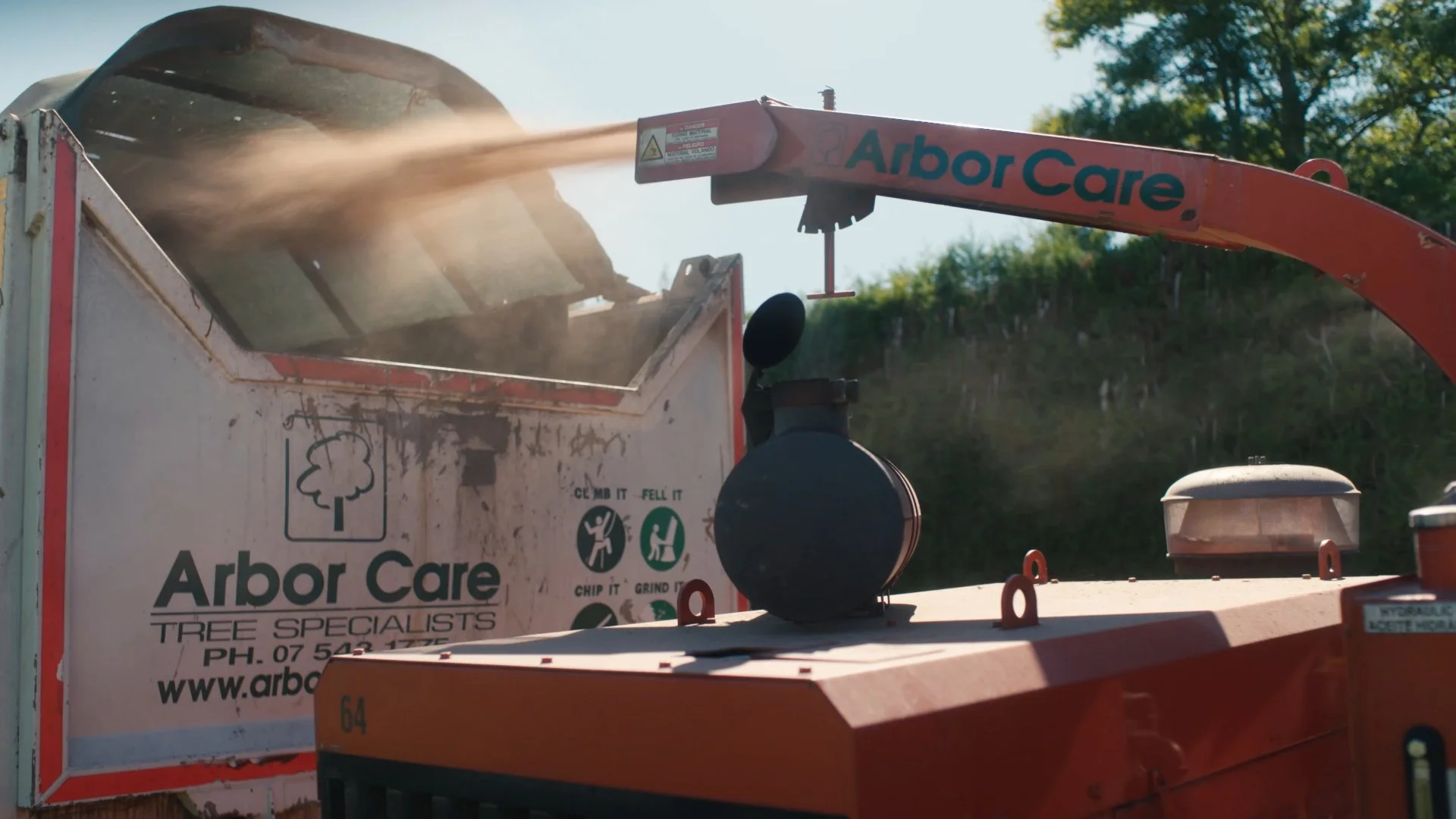 A wood chipper in use with wood chips being discharged into a truck with Arbor Care Tree Specialists branding, outdoors on a sunny day.