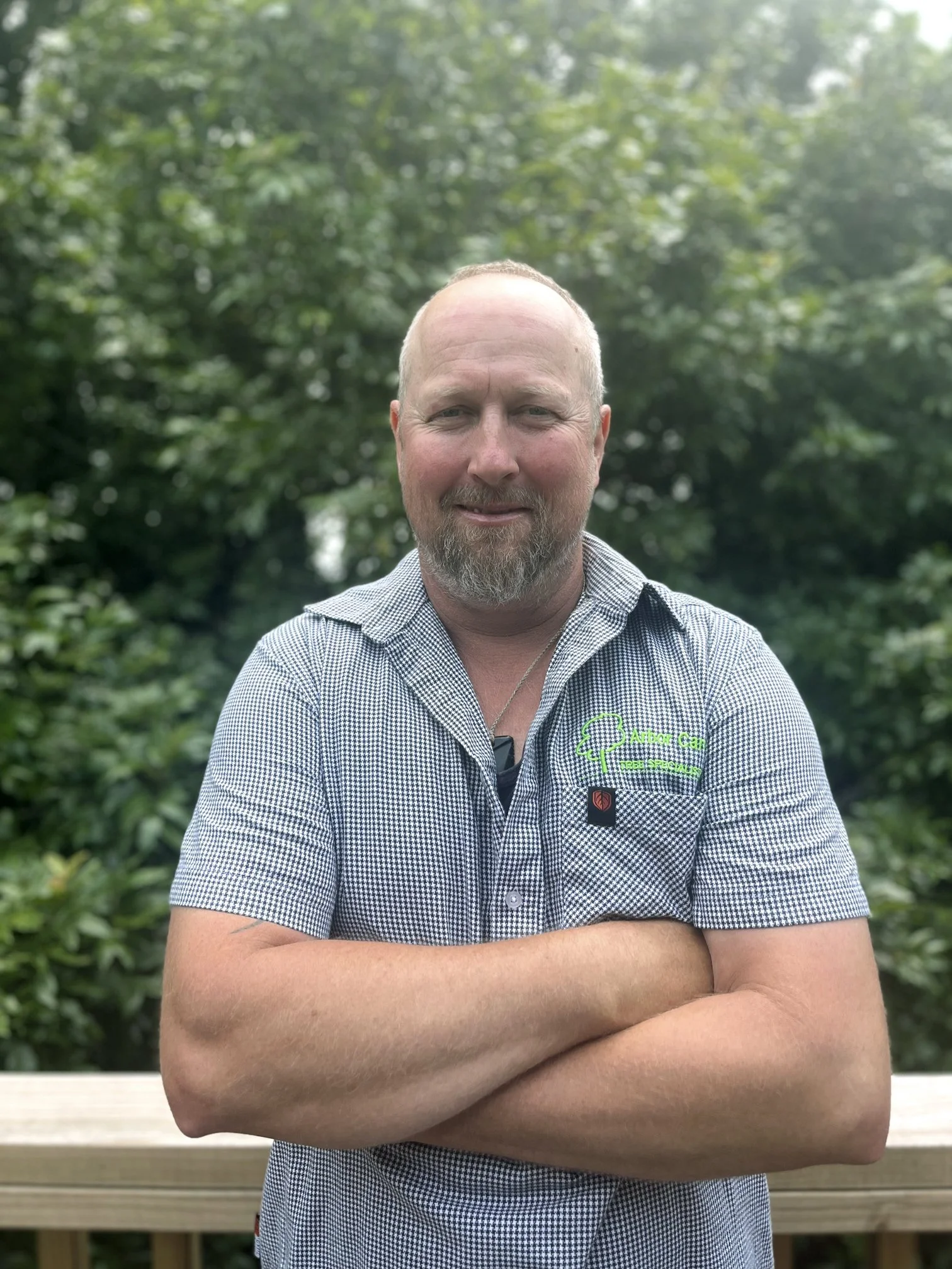 A man with a beard and short hair standing outdoors with arms crossed, wearing a checkered shirt with a logo on the left chest that says 'Aaron Green Tree Service,' in front of green trees and bushes.