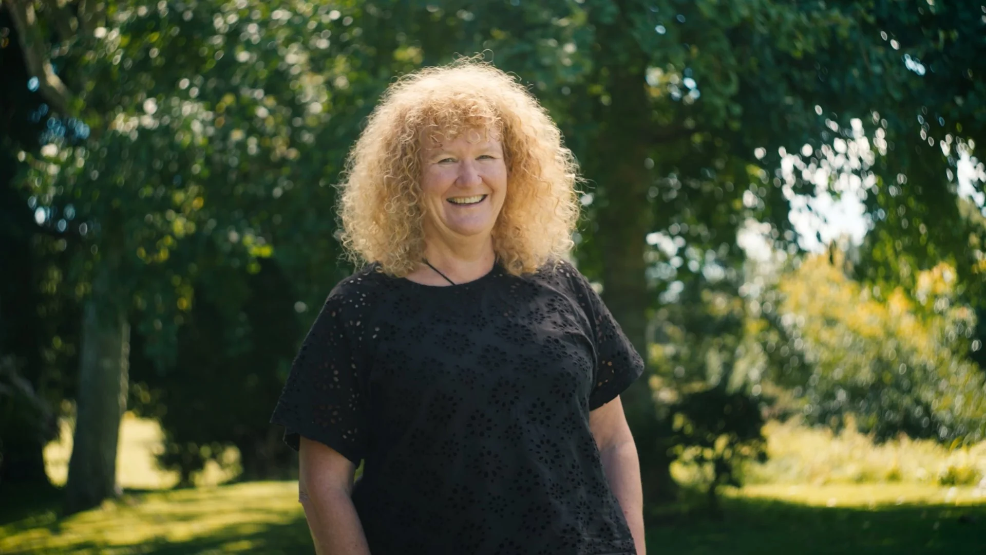 A smiling woman with curly blonde hair wearing a black top outdoors with trees and sunlight in the background.