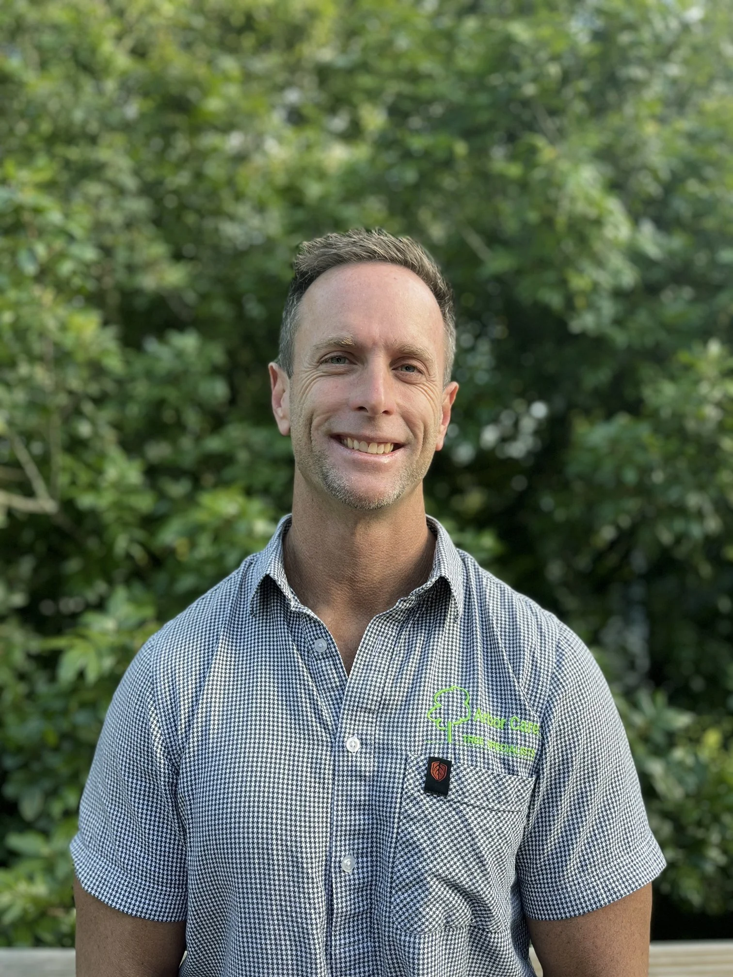 A man smiling outdoors with a background of green trees, wearing a checkered shirt with a logo that reads 'Andi Coffee Tree Scientist'.