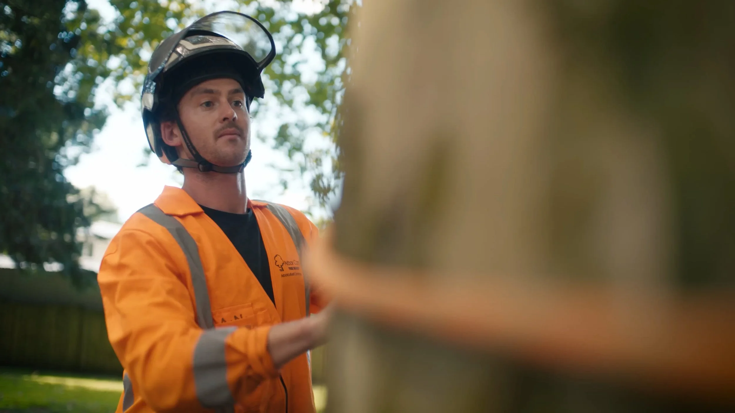 A man wearing an orange safety jacket and a safety helmet with a face shield, standing outdoors next to a wooden fence, with trees and sunlight in the background.