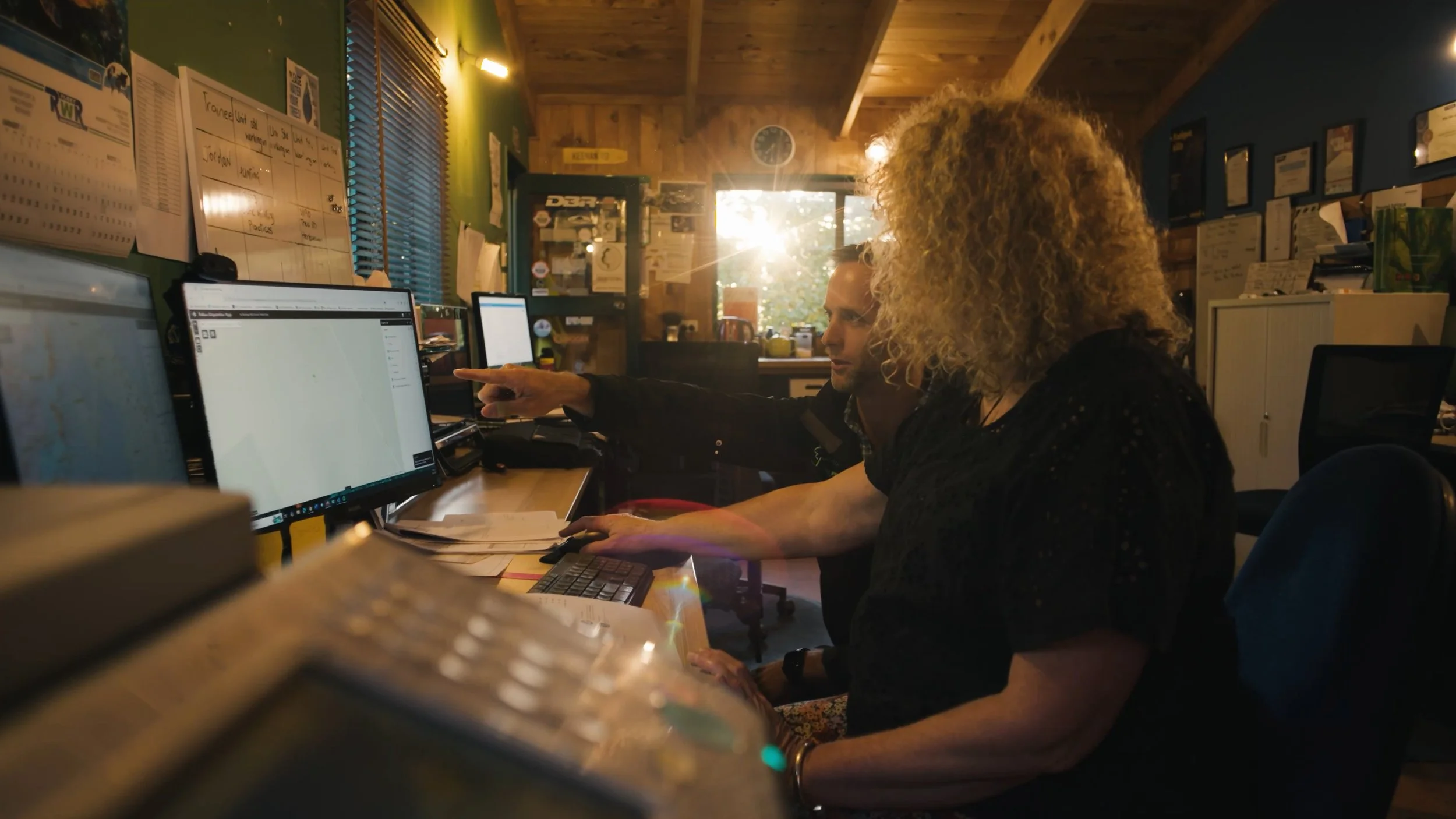 Two people working at a desk in an office, looking at computer screens, with sunlight coming through a window behind them.