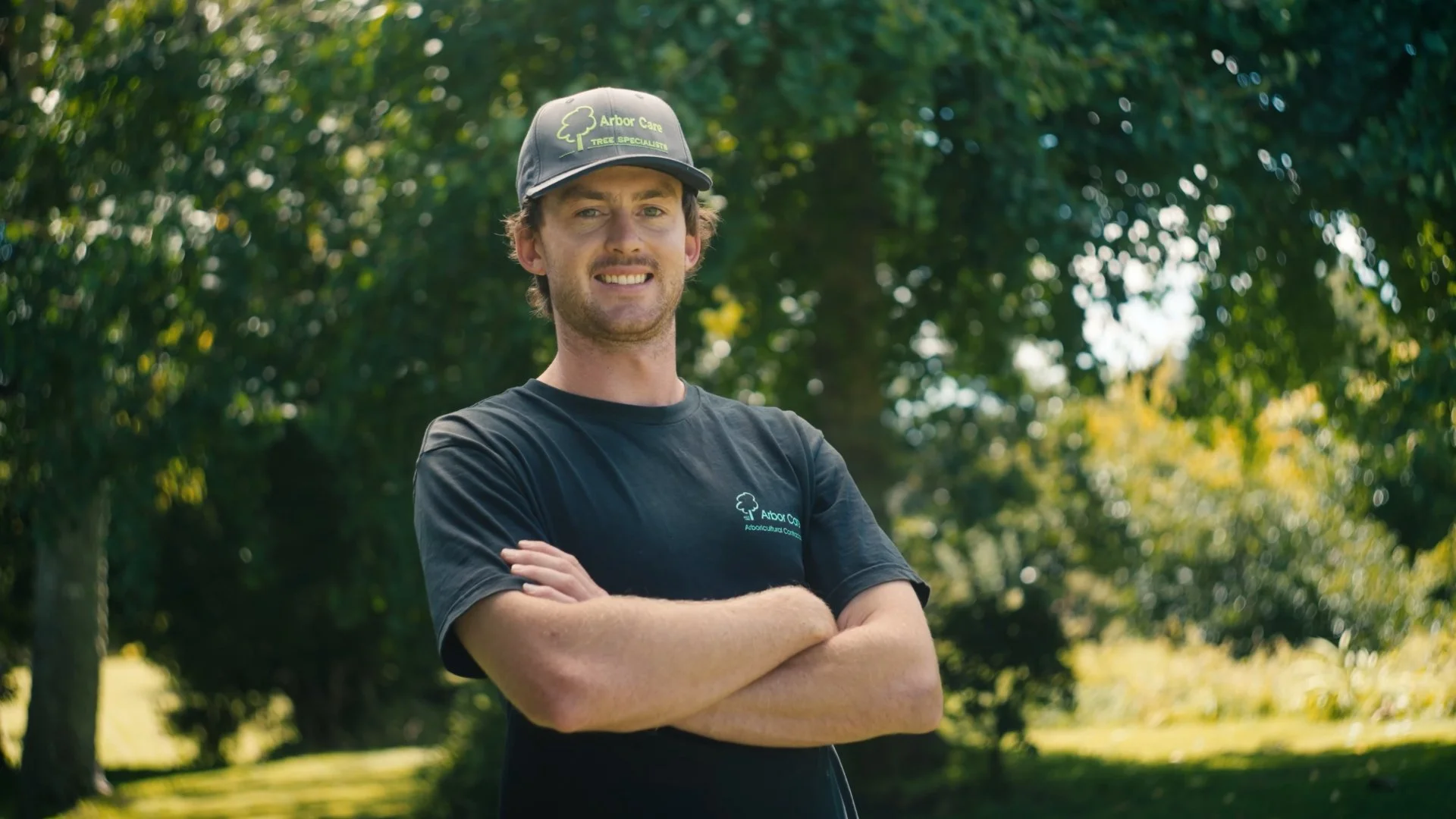 A man standing outdoors among trees, wearing a black t-shirt and a cap with the logo and text for Arbor Care. He is smiling and has his arms crossed.