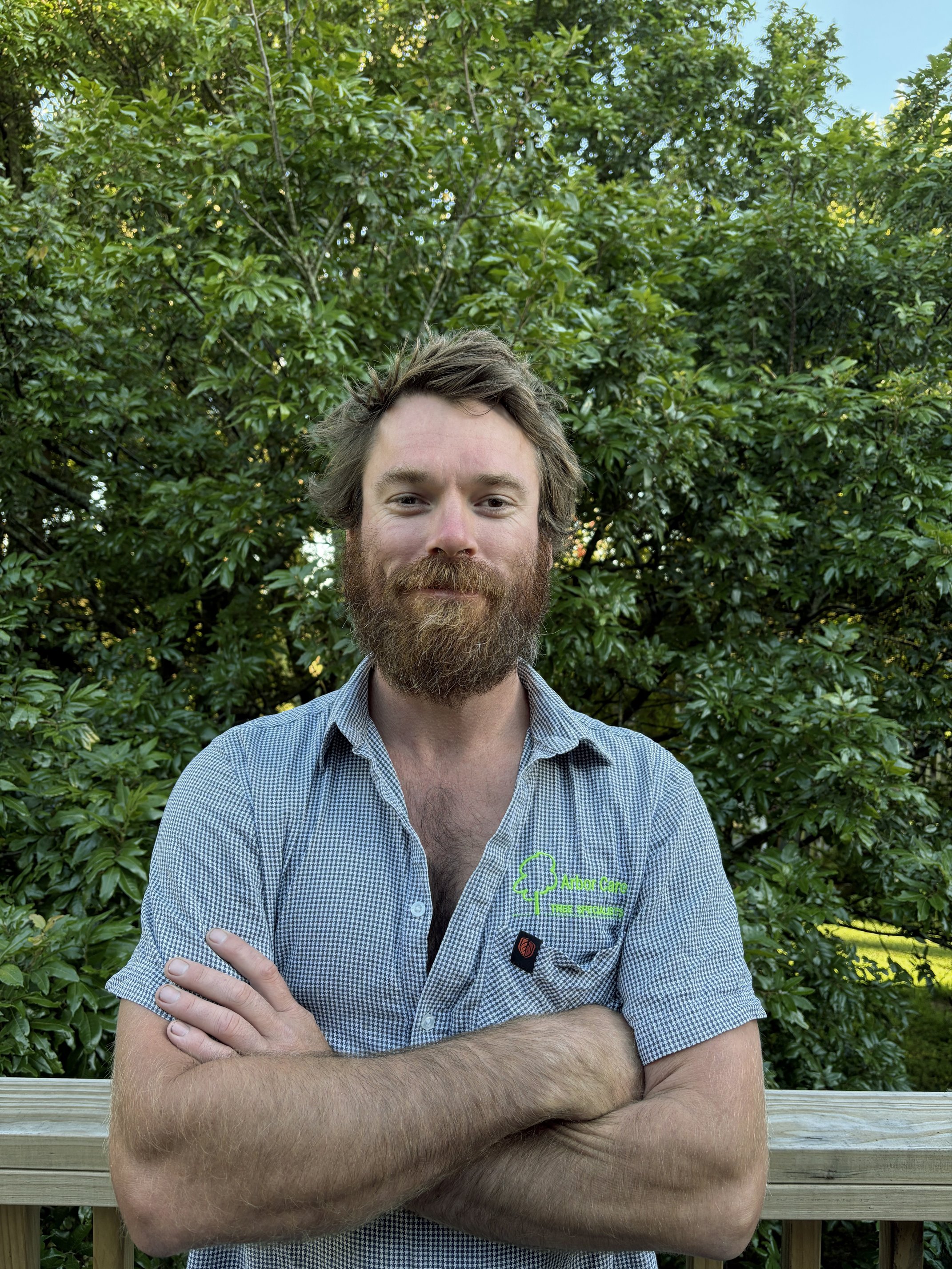 A man with a beard and mustache standing outdoors in front of a leafy green tree, crossing his arms and looking at the camera, wearing a checkered short-sleeve shirt with a logo on the chest.
