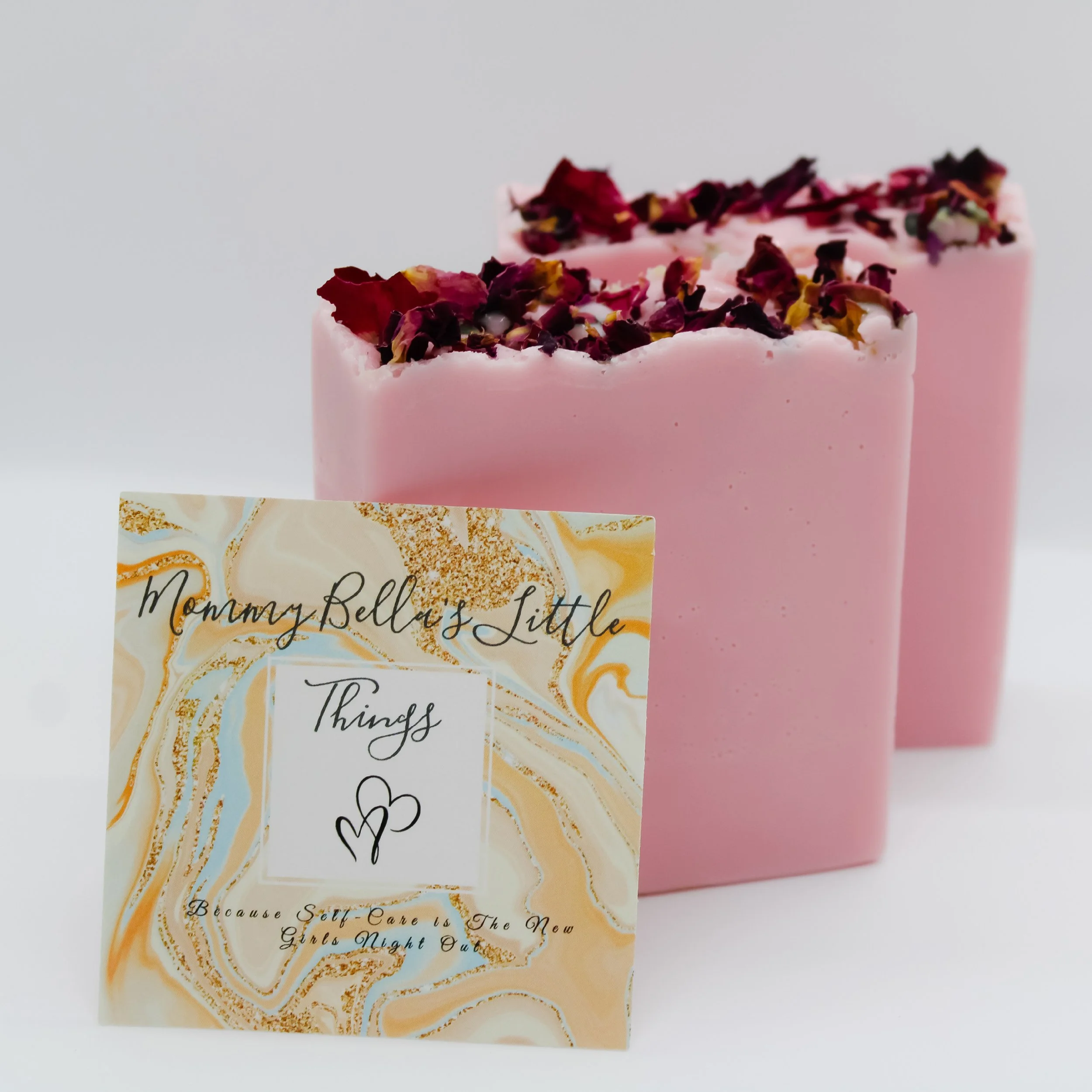 Two pink soap bars topped with dried rose petals, with a card in front reading "Mommy Bell's Little Things" and a subtitle "Because Self-Care is the New Girls Night Out."