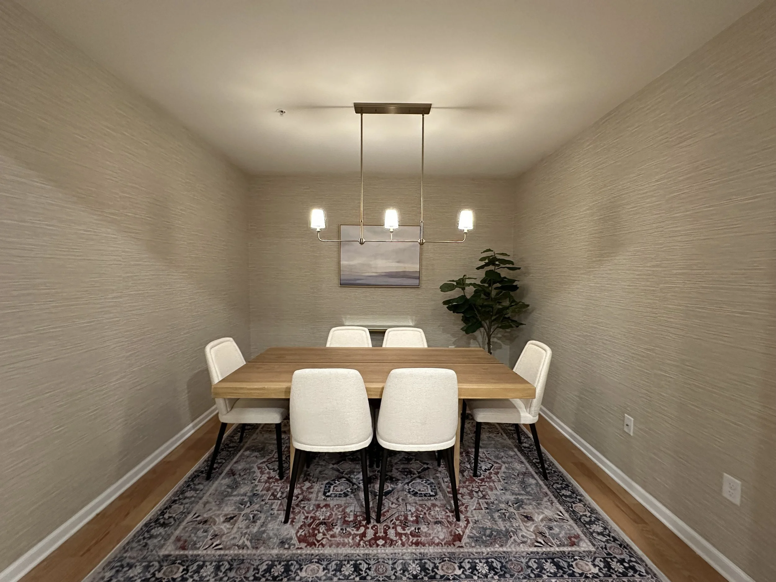 Empty dining room with a wooden table, six white upholstered chairs, a chandelier with four lights, a framed landscape on the wall, a potted plant, and an area rug on hardwood flooring.