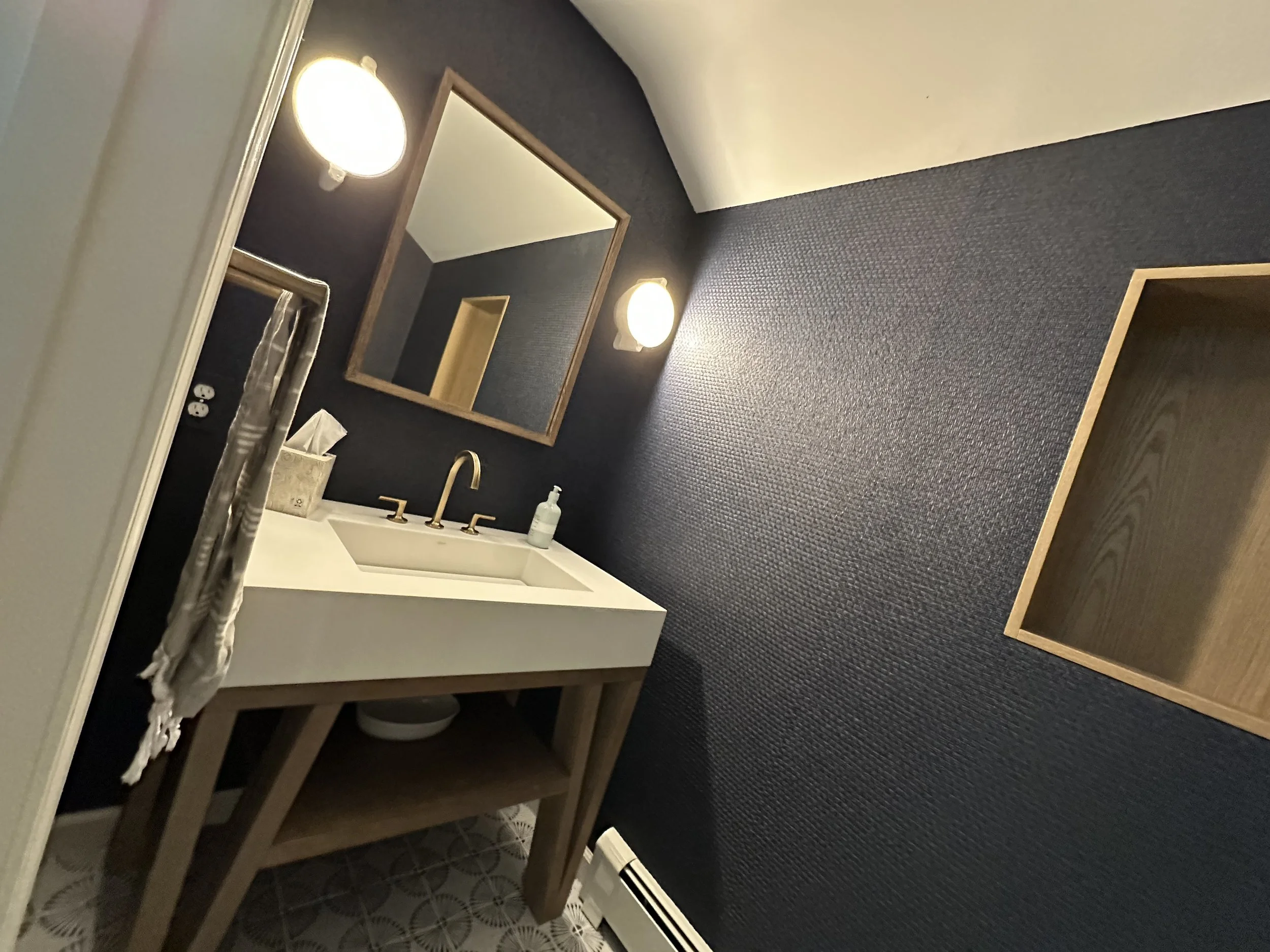 Bathroom with dark textured wall, wooden framed mirror, white sink with bronze faucet, soap dispenser, and open wooden shelf, illuminated by two round wall-mounted lights.