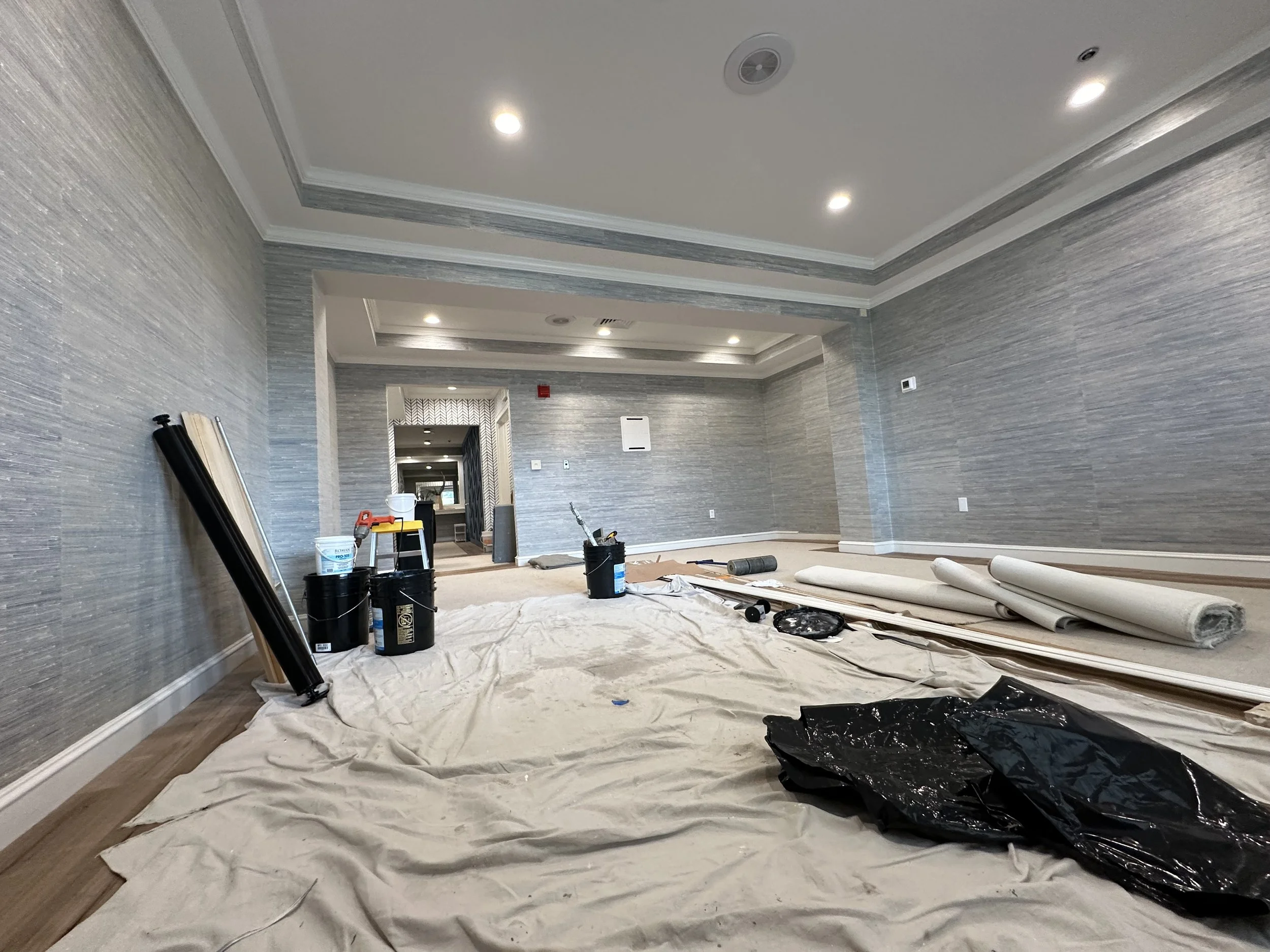 Room under renovation with painting supplies, rolls of wallpaper, and painter's tarp on the floor, featuring gray textured walls and ceiling lights.