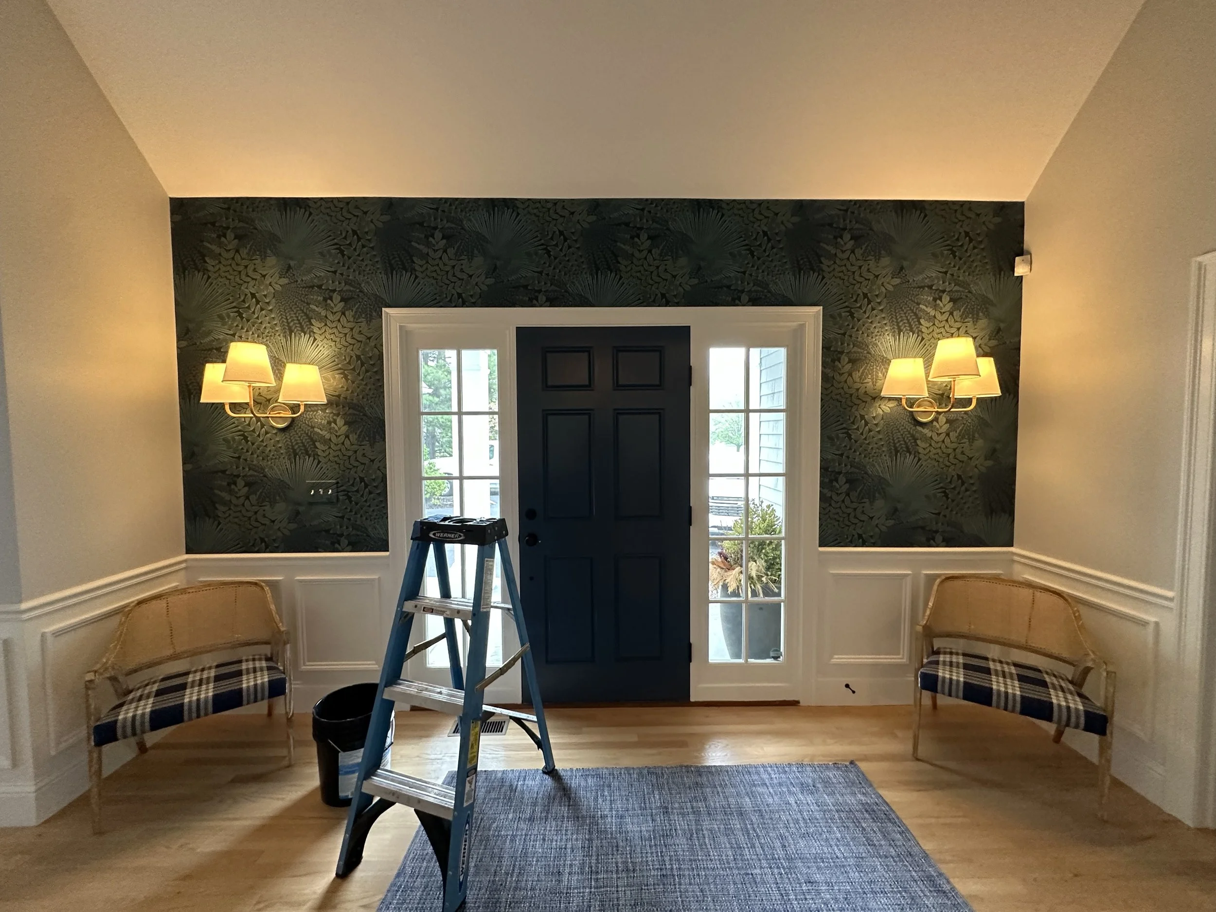 Foyer area with dark front door, sidelights, two wall sconces, two chairs against walls, ladder and bucket on wooden floor, and a gray rug.