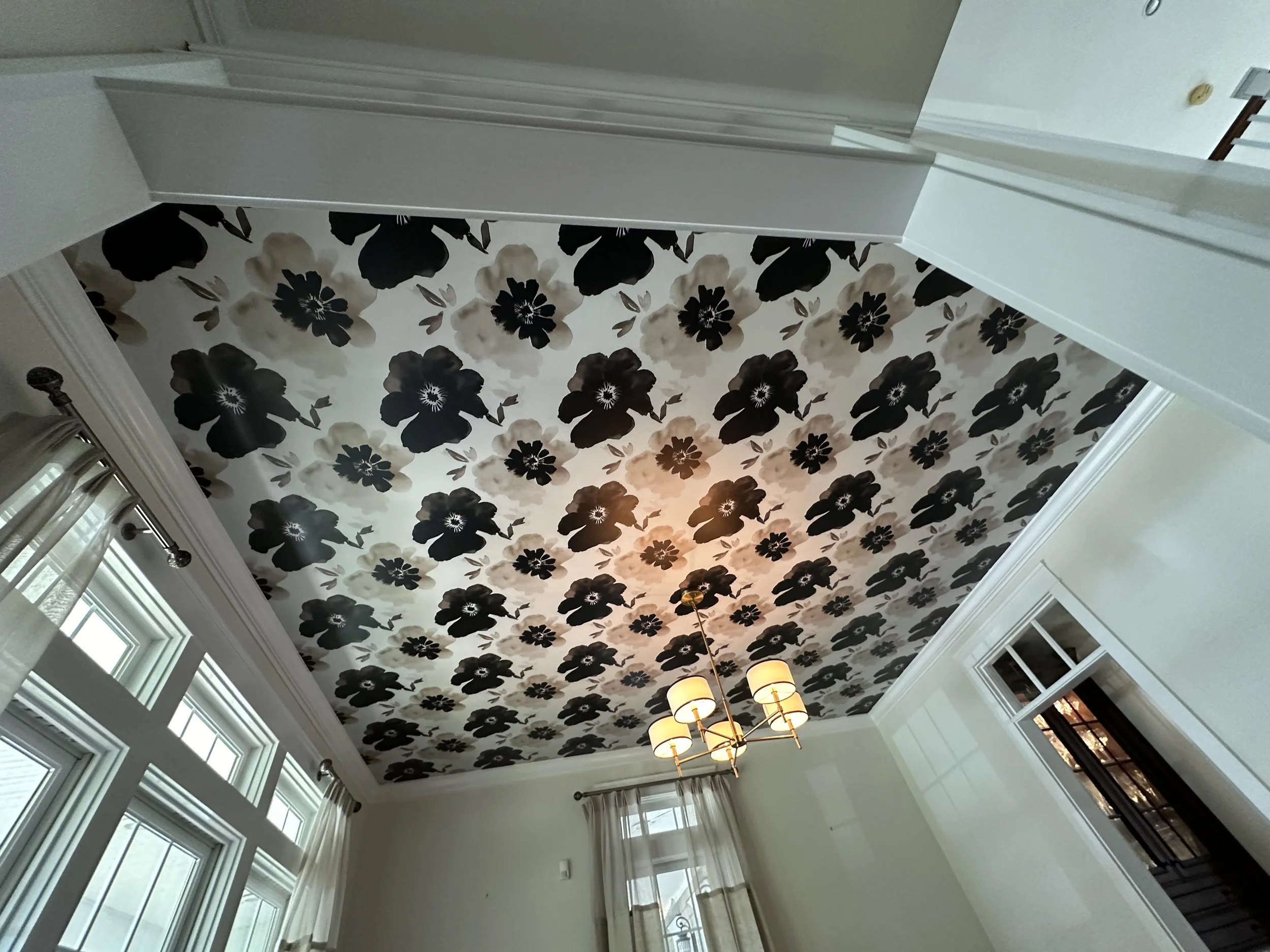 Room ceiling with black and white floral wallpaper, chandelier with five lampshades, large windows with white curtains.