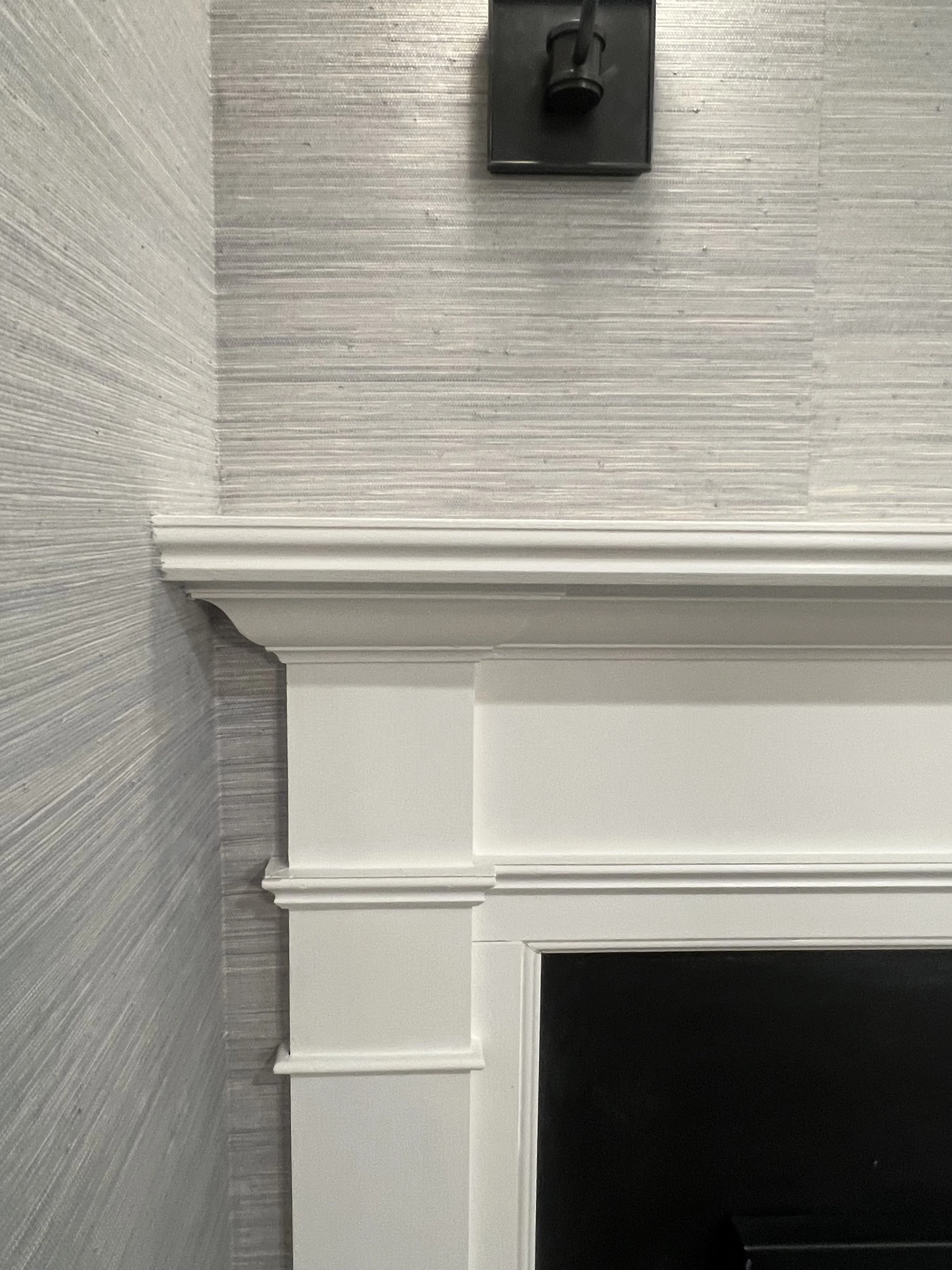 Close-up of a white fireplace mantel with crown molding against a textured gray wallpaper wall, including an electrical outlet and a mounted dark-colored device above the mantel.