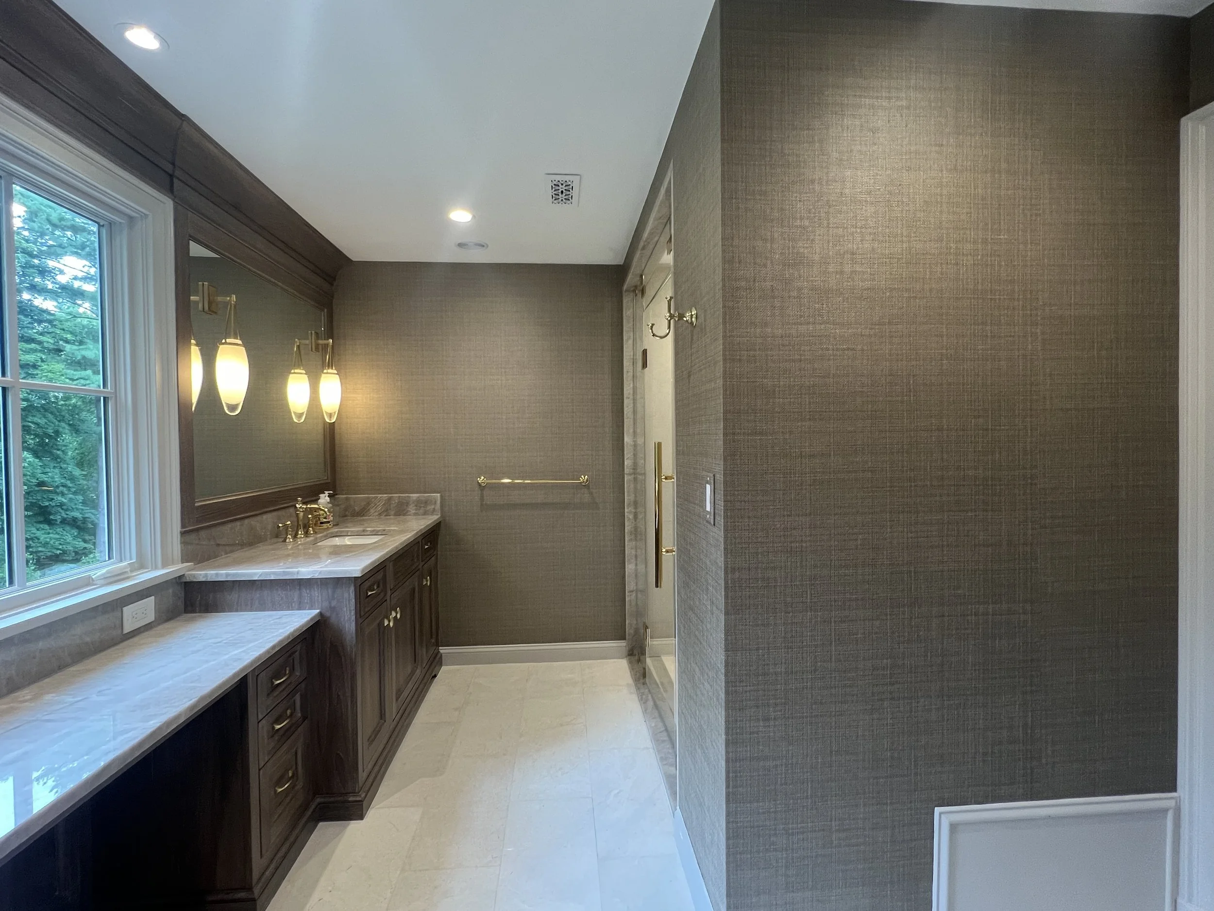 Bathroom with a large window, dark wood vanity with marble countertop, gold fixtures, and beige textured wallpaper.