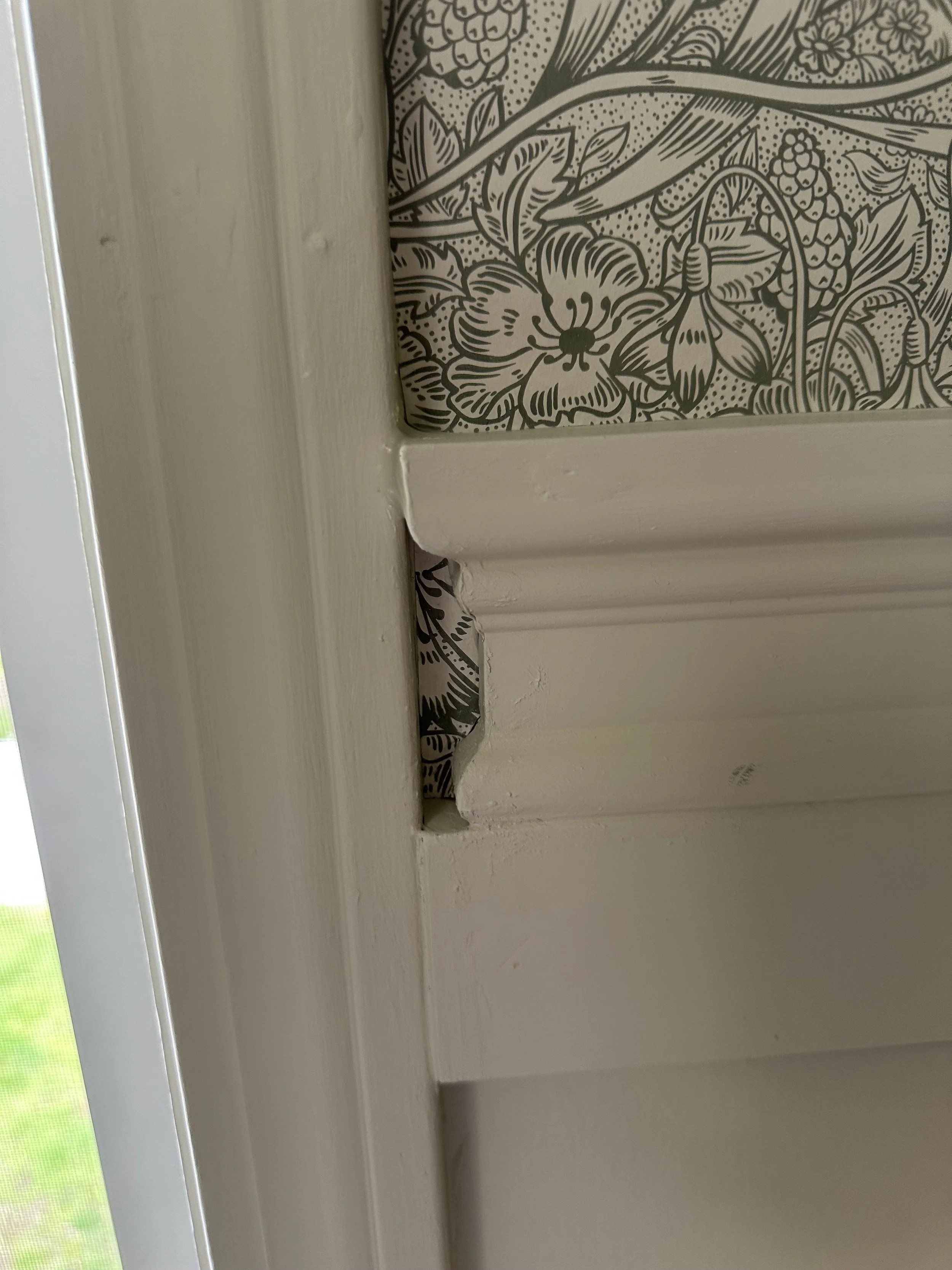 Close-up of a section of a wall with wallpaper featuring floral and botanical patterns behind a white window frame with damage at the bottom.