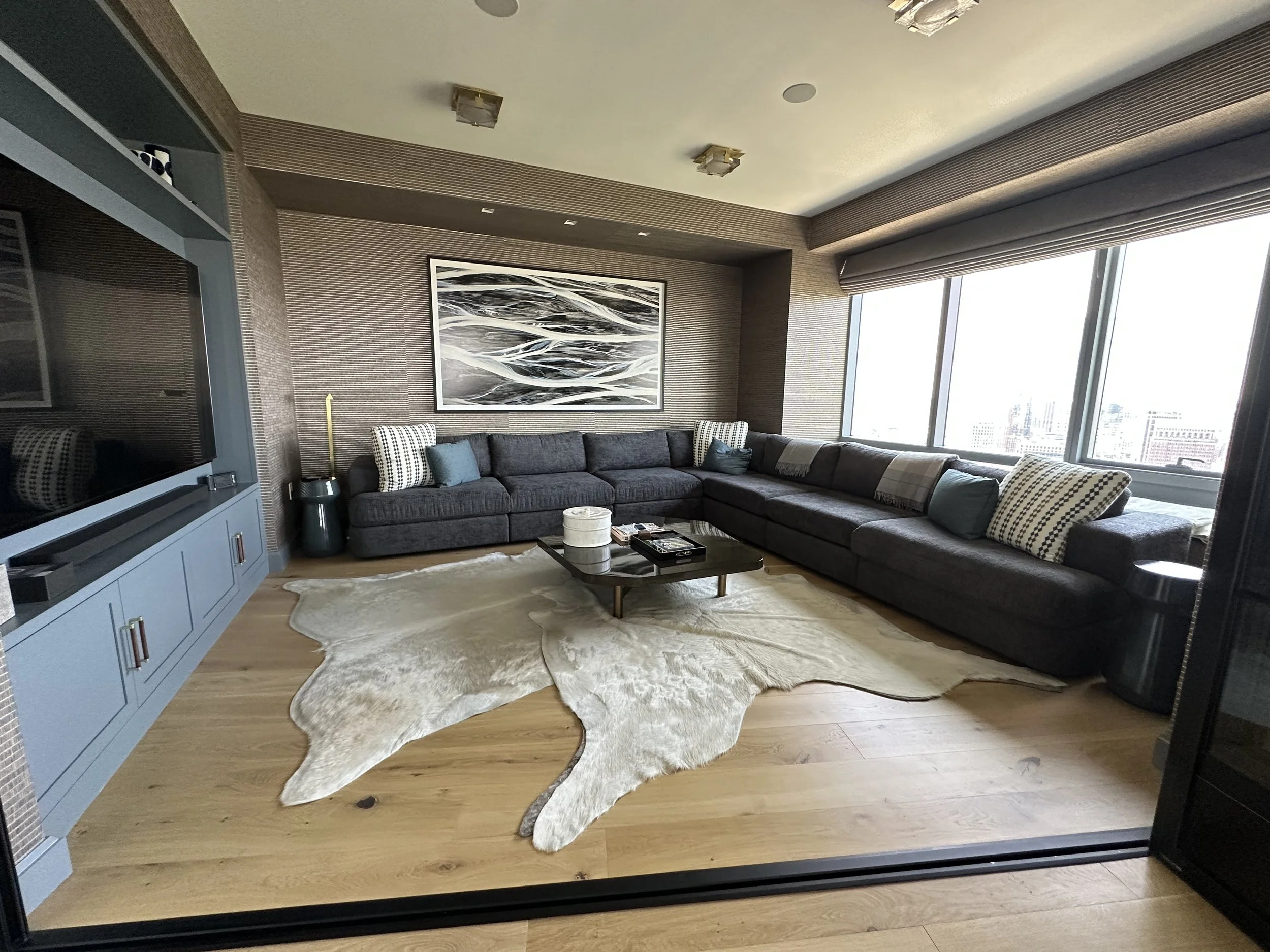 Living room with a large L-shaped dark gray sofa, a wooden coffee table, a textured wall with modern art, large windows with city view, and a cowhide rug on light hardwood floors.