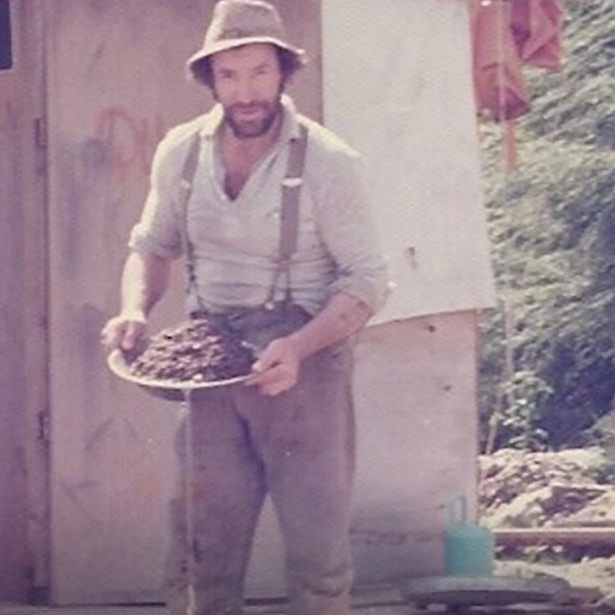 A man wearing a hat, suspenders, and a light-colored shirt, holding a tray of what appears to be a dark dish, standing outdoors beside a building, with trees in the background.