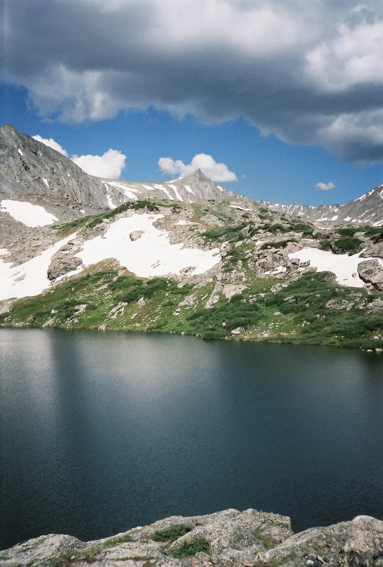 Mountain lake with rocky shores, snow patches on green mountains, partly cloudy sky.
