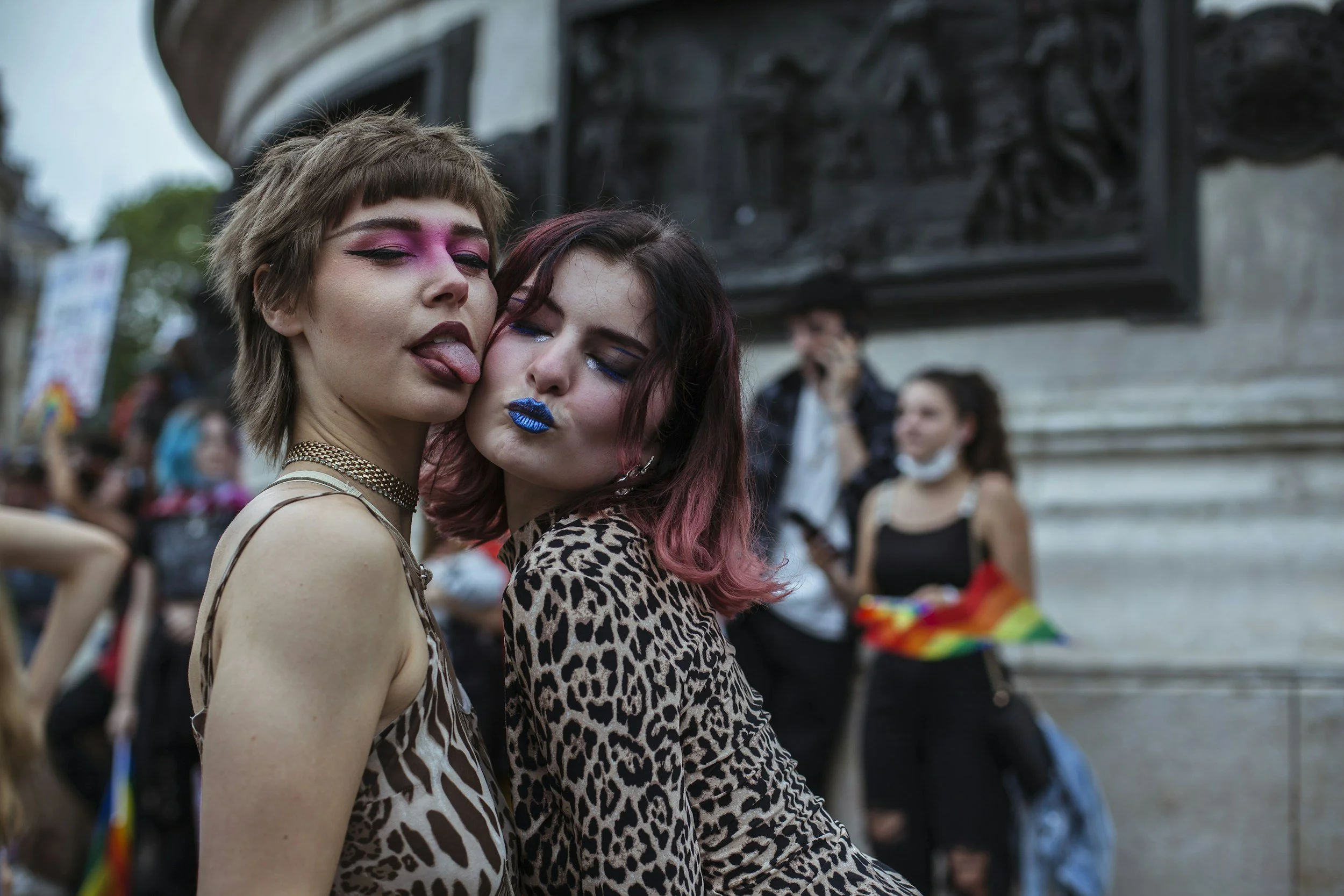 Two young queer people with colorful makeup and animal print clothing at a pride event, posing closely together with a crowd in the background.
