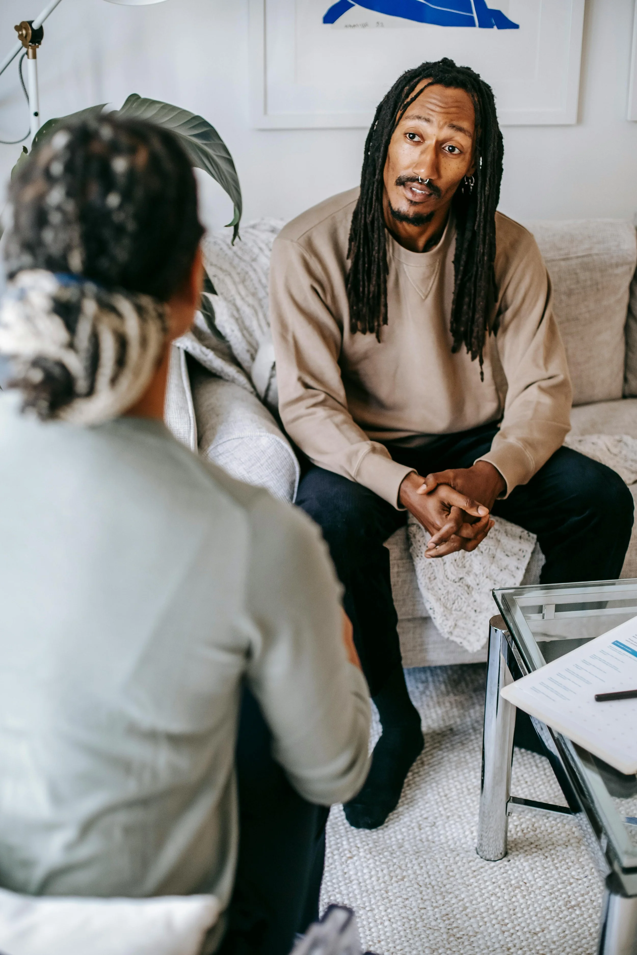 A man with long dreadlocks and a beard sitting on a beige couch, talking to a woman with braids, in a living room with artwork on the wall.