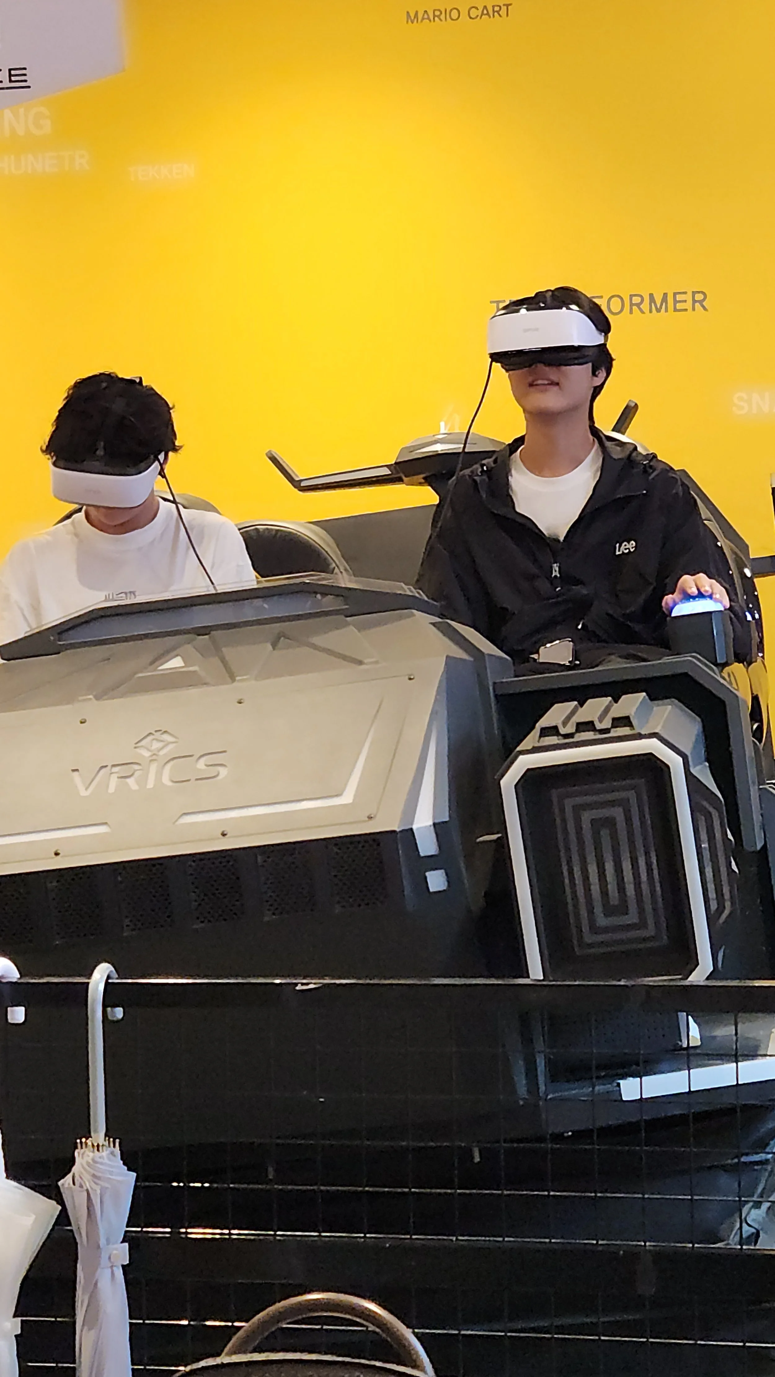 Two young men wearing virtual reality headsets and seated in a VR racing simulator with a yellow background and various gaming slogans.