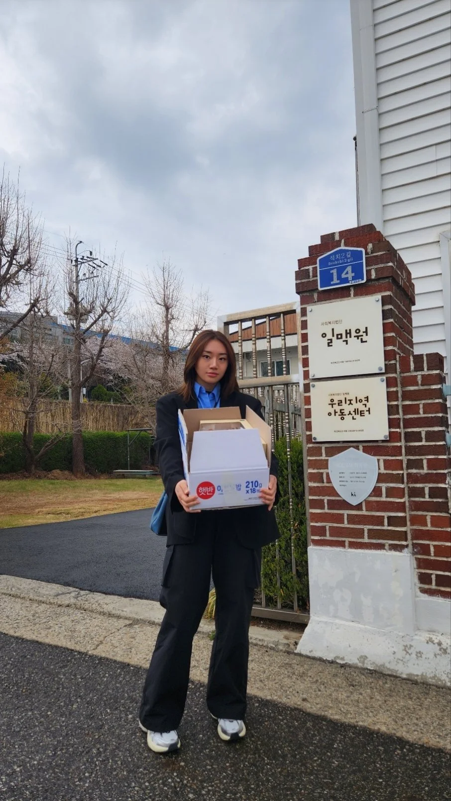 A young woman in a black suit and sneakers holding a box of food standing outside next to a brick sign with blue street number 14, in front of a white building with trees and cloudy sky in the background.