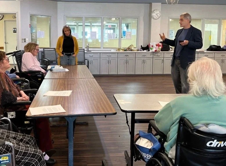 A group of people in a room, some in wheelchairs, listening to a man speaking at the front. The room has a long table, cabinets, large windows, and a clock on the wall.