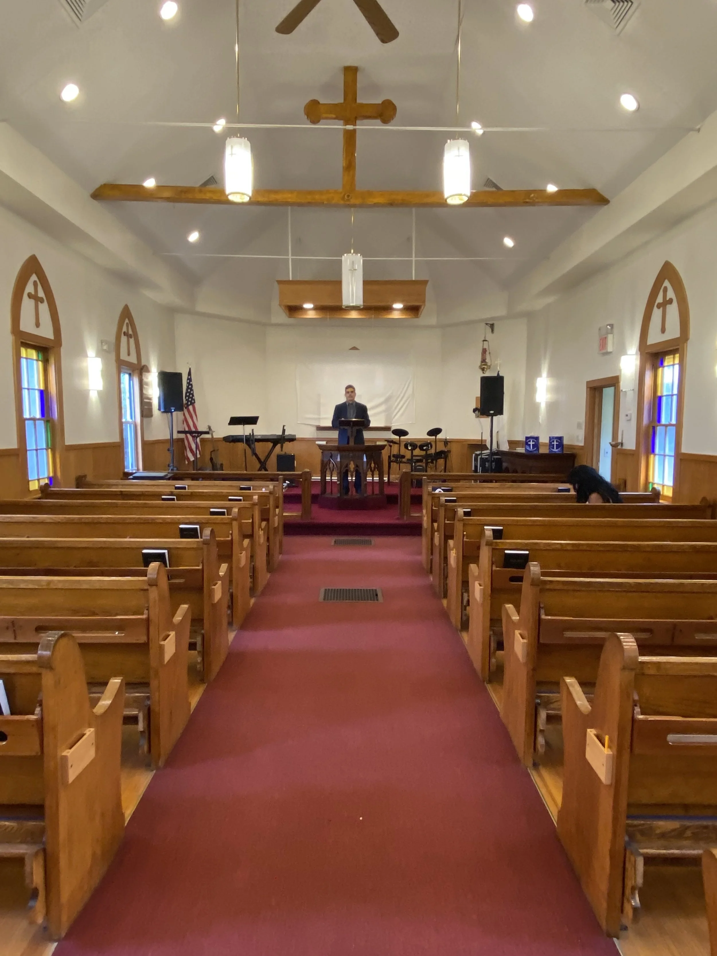 Interior of a church with wooden pews, stained glass windows, a cross hanging from the ceiling, and a person standing at the pulpit at the front.