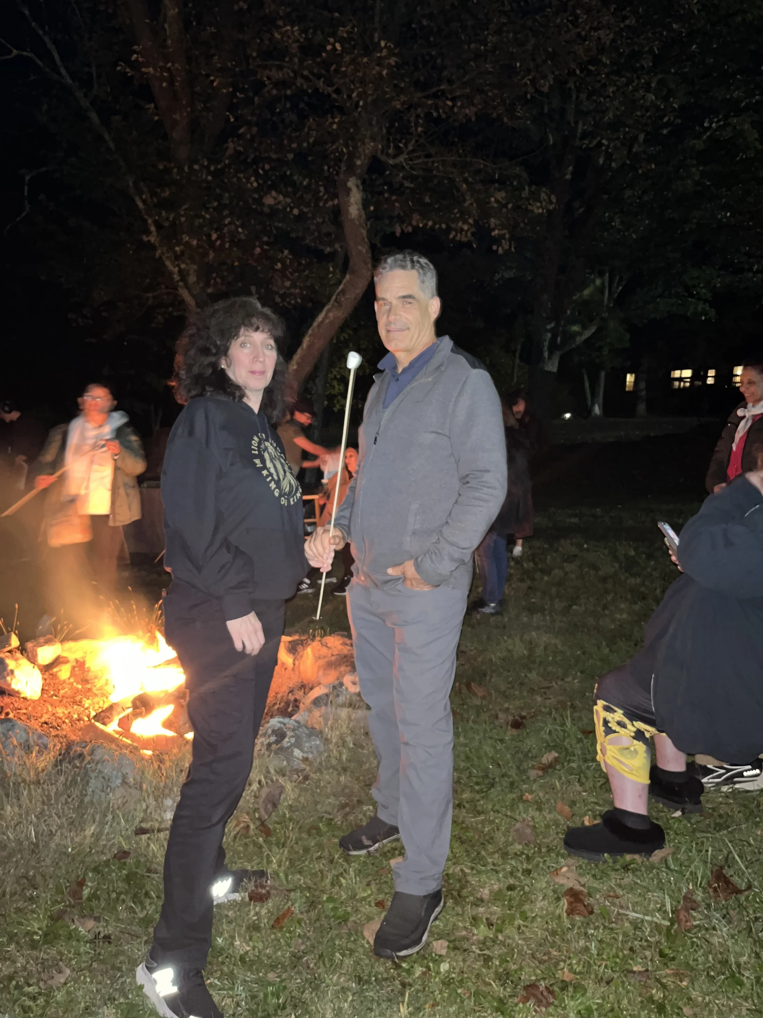 A man and woman standing outdoors at night near a campfire, with trees in the background. The woman is holding a marshmallow on a stick, and other people are visible around the fire, some sitting or standing, some on their phones.