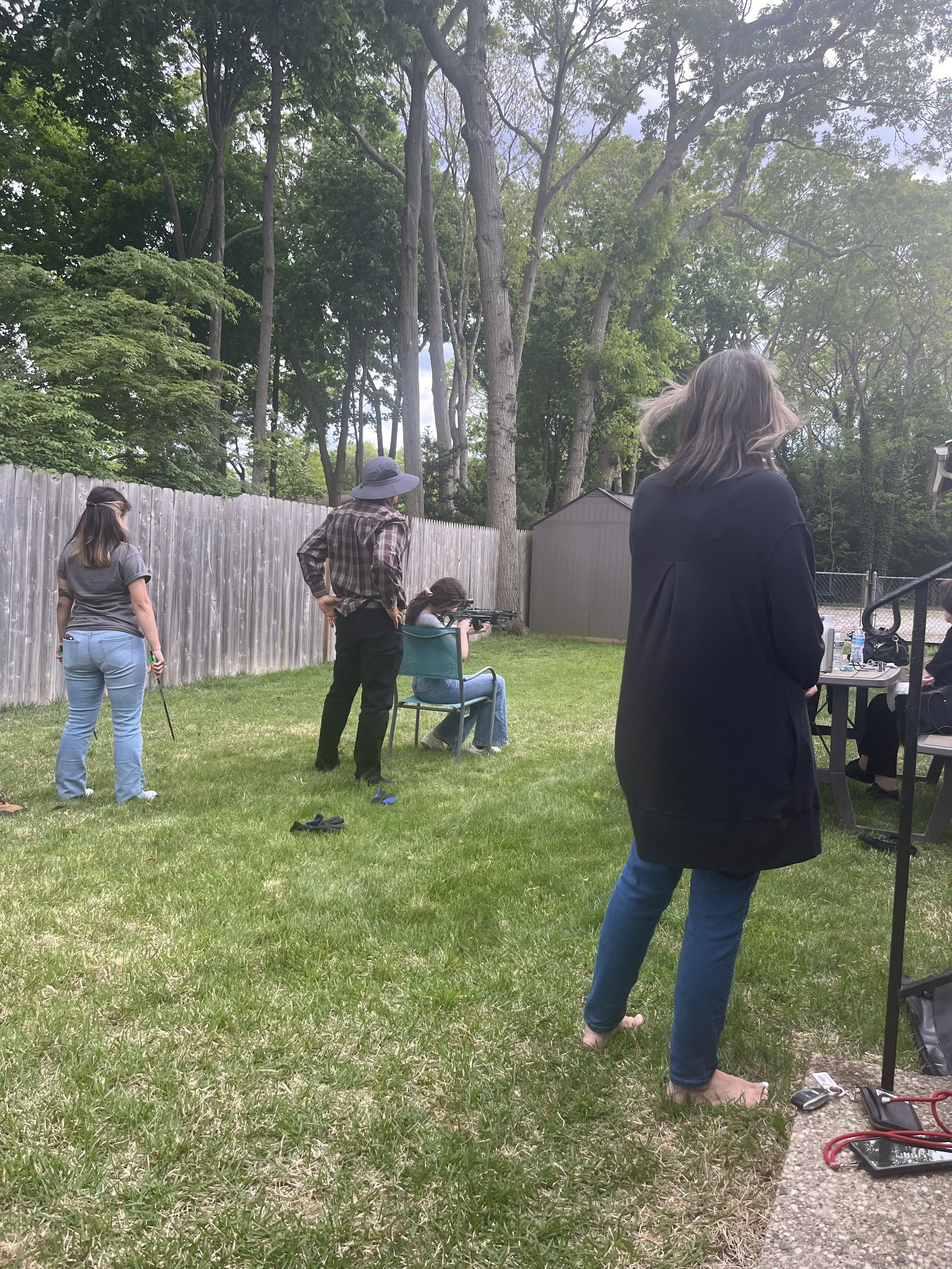 Several people outdoors in a backyard, with one woman wearing a hat and plaid shirt aiming a rifle, others standing or sitting nearby, trees in the background, and a weather-shielded shed.