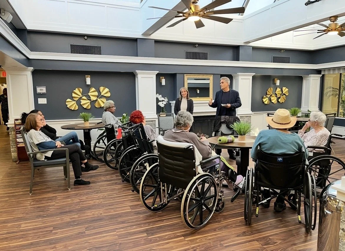 Group of elderly people seated in wheelchairs and chairs, listening to a man speaking and a woman standing beside him in a well-lit indoor lounge with dark blue walls and gold decorations.