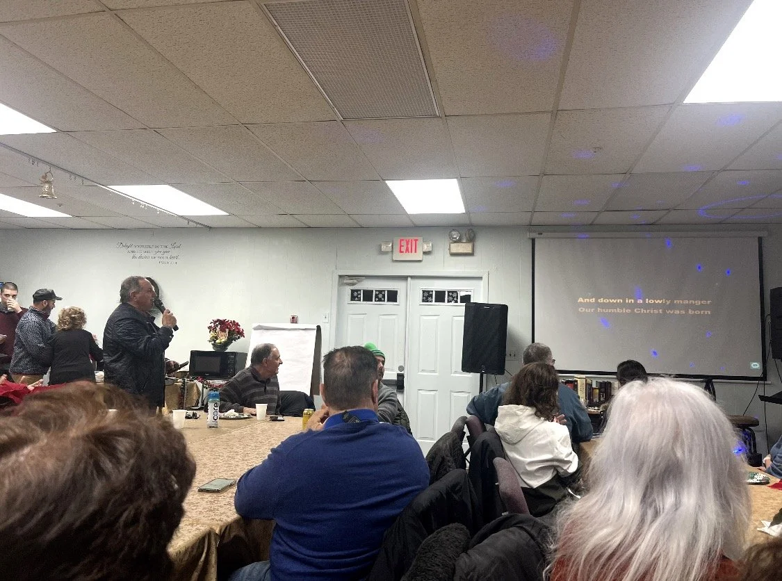 People attending a meeting or presentation in a room with a speaker standing near a table and a projection screen displaying song lyrics.