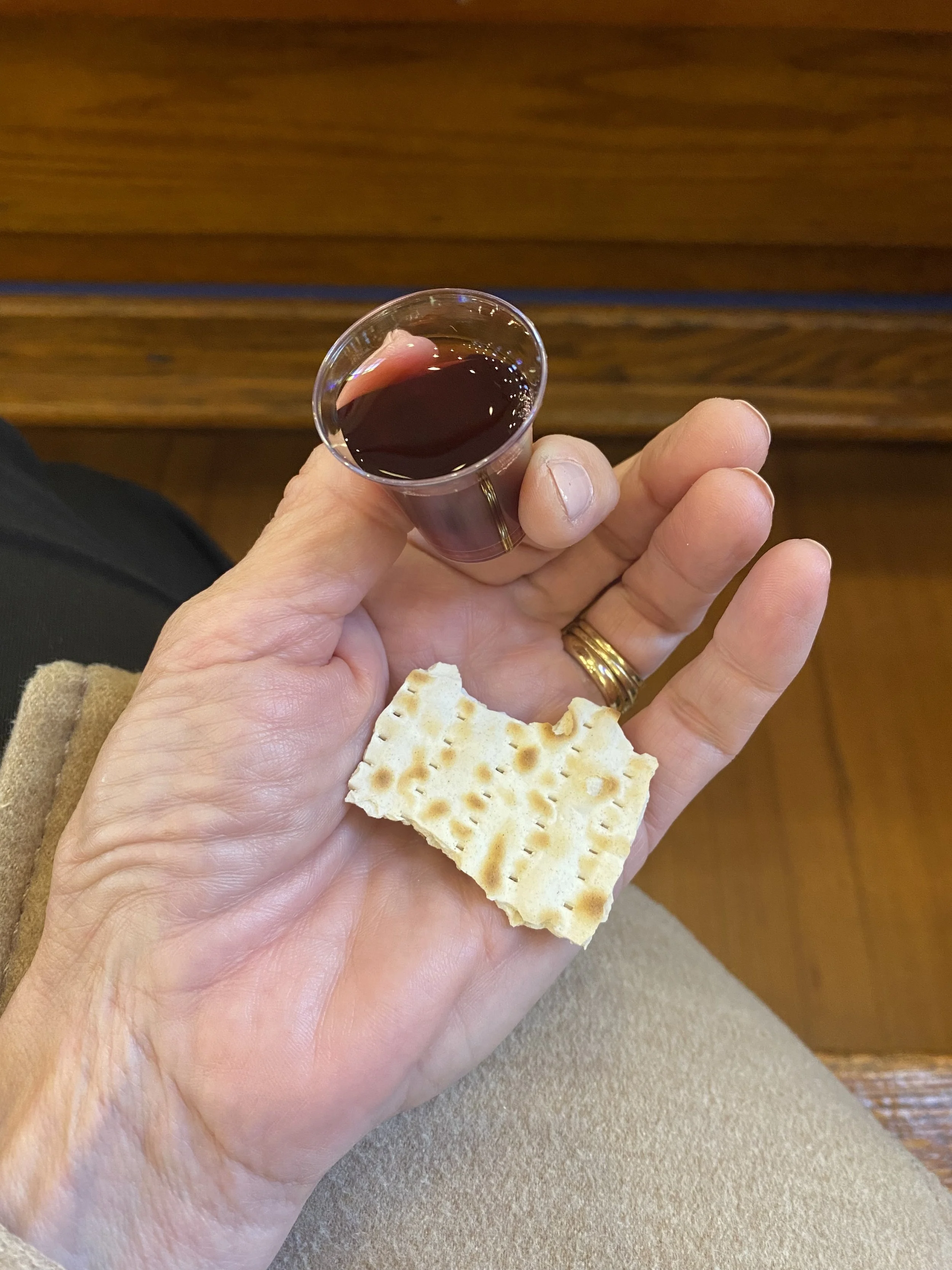 A person's hand holding a small glass of dark red wine and a piece of saltine cracker. The person is wearing a gold ring and is dressed in beige. The background shows a wooden surface.
