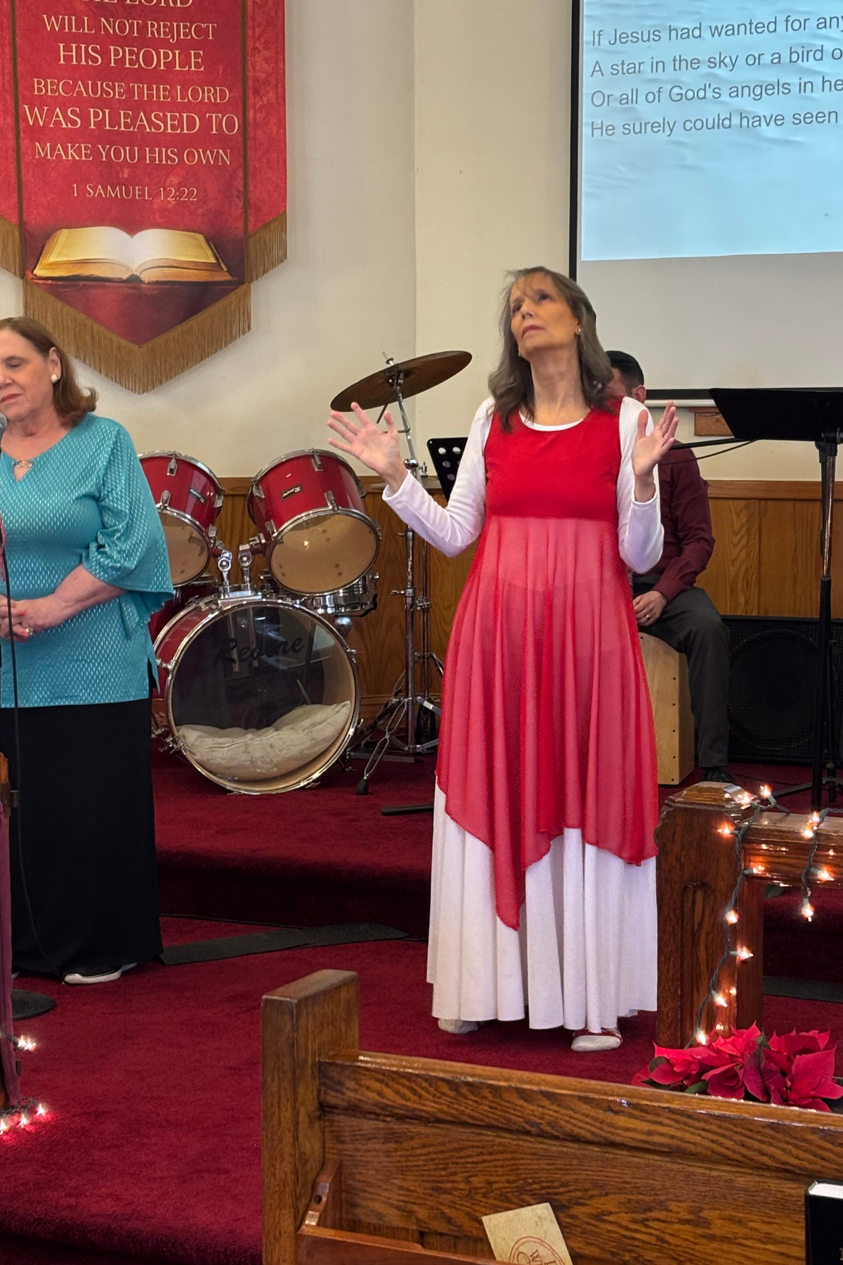 A woman in a red and white dress is standing on a stage with her hands raised, possibly singing or speaking, with a drum set and other musicians behind her. There is a red banner with gold fringes and a Bible verse on the wall, and a large screen with text projected on it.